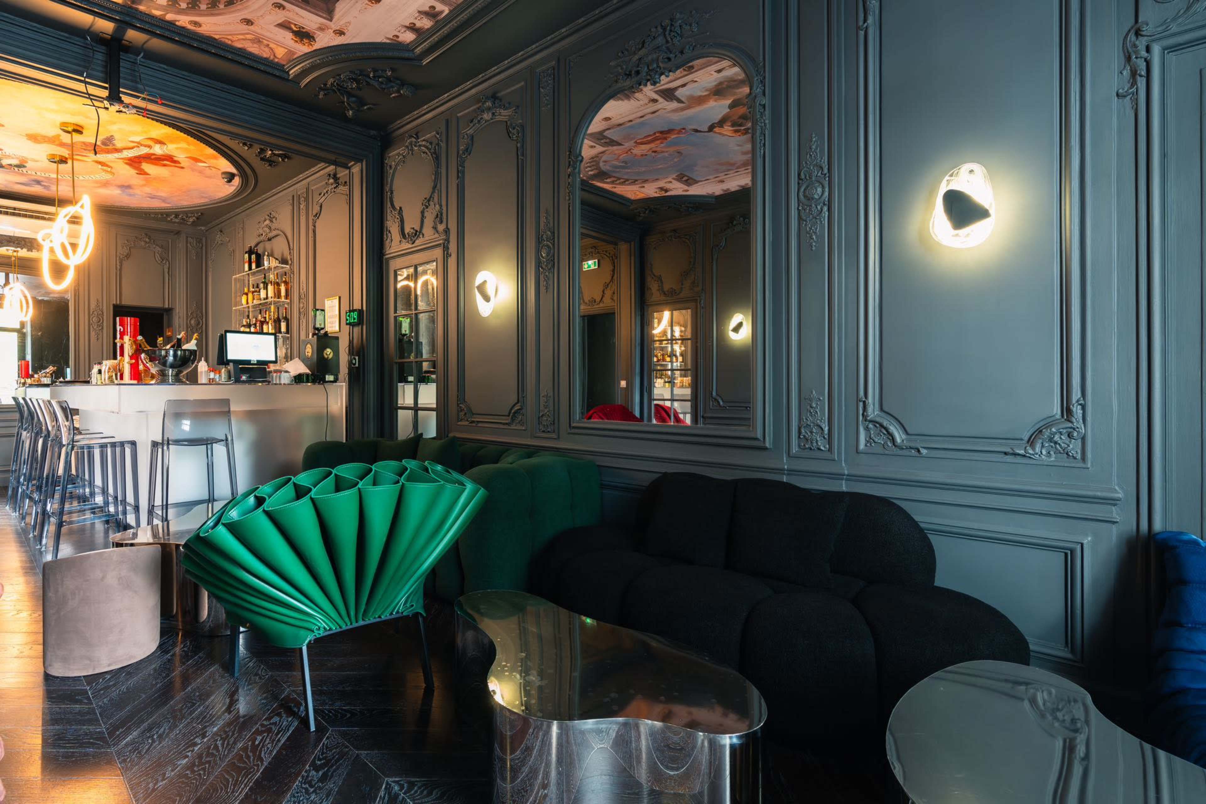 The image shows a stylish lounge area featuring a bar with a modern design, ornate wall decor, and plush seating arrangements in various colors.