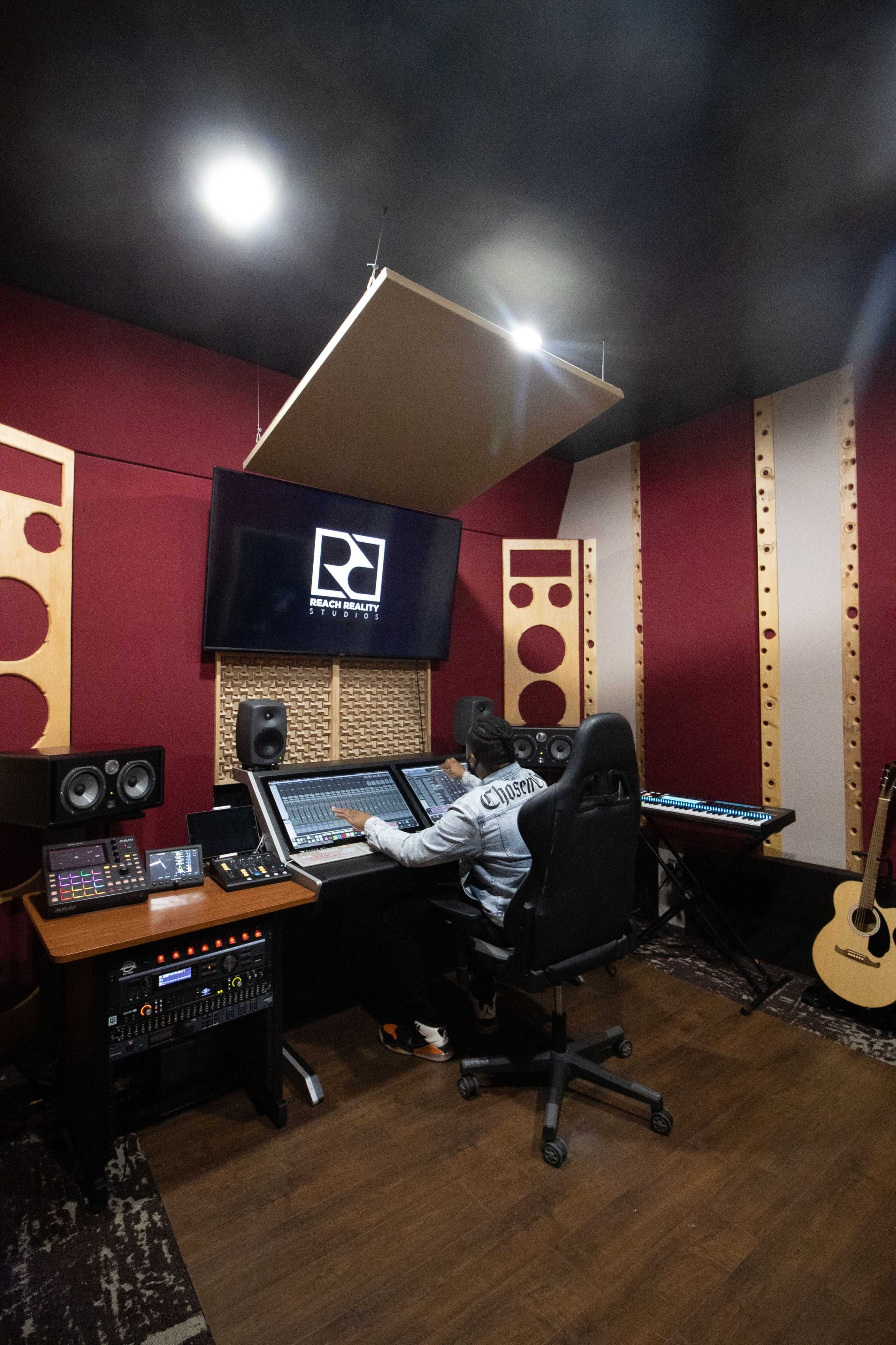 A person sits at a mixing console in a modern recording studio, surrounded by audio equipment and instruments.