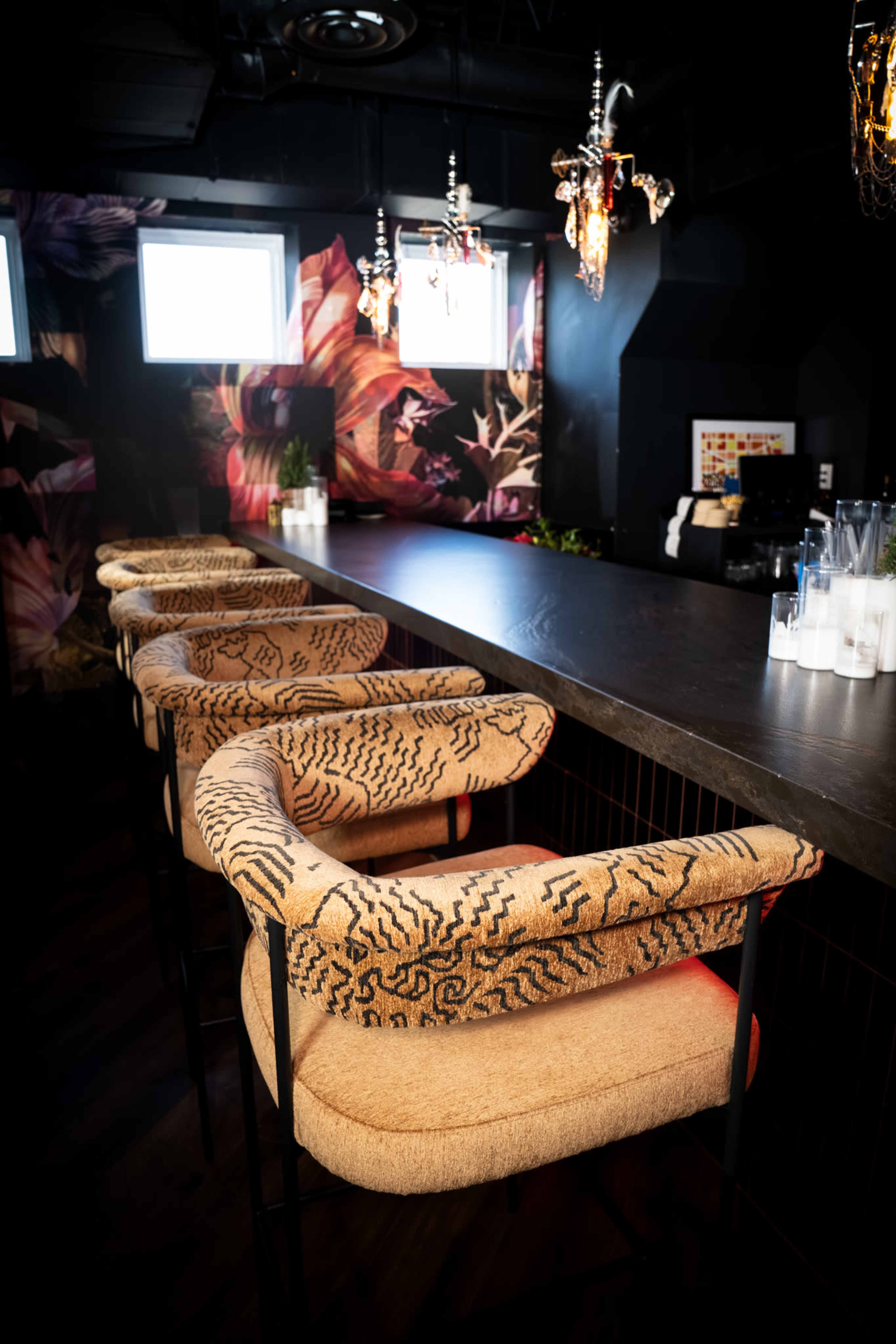 The image shows a stylish bar area featuring four brown and patterned high chairs along a sleek black countertop, with decorative floral wallpaper and elegant light fixtures overhead.
