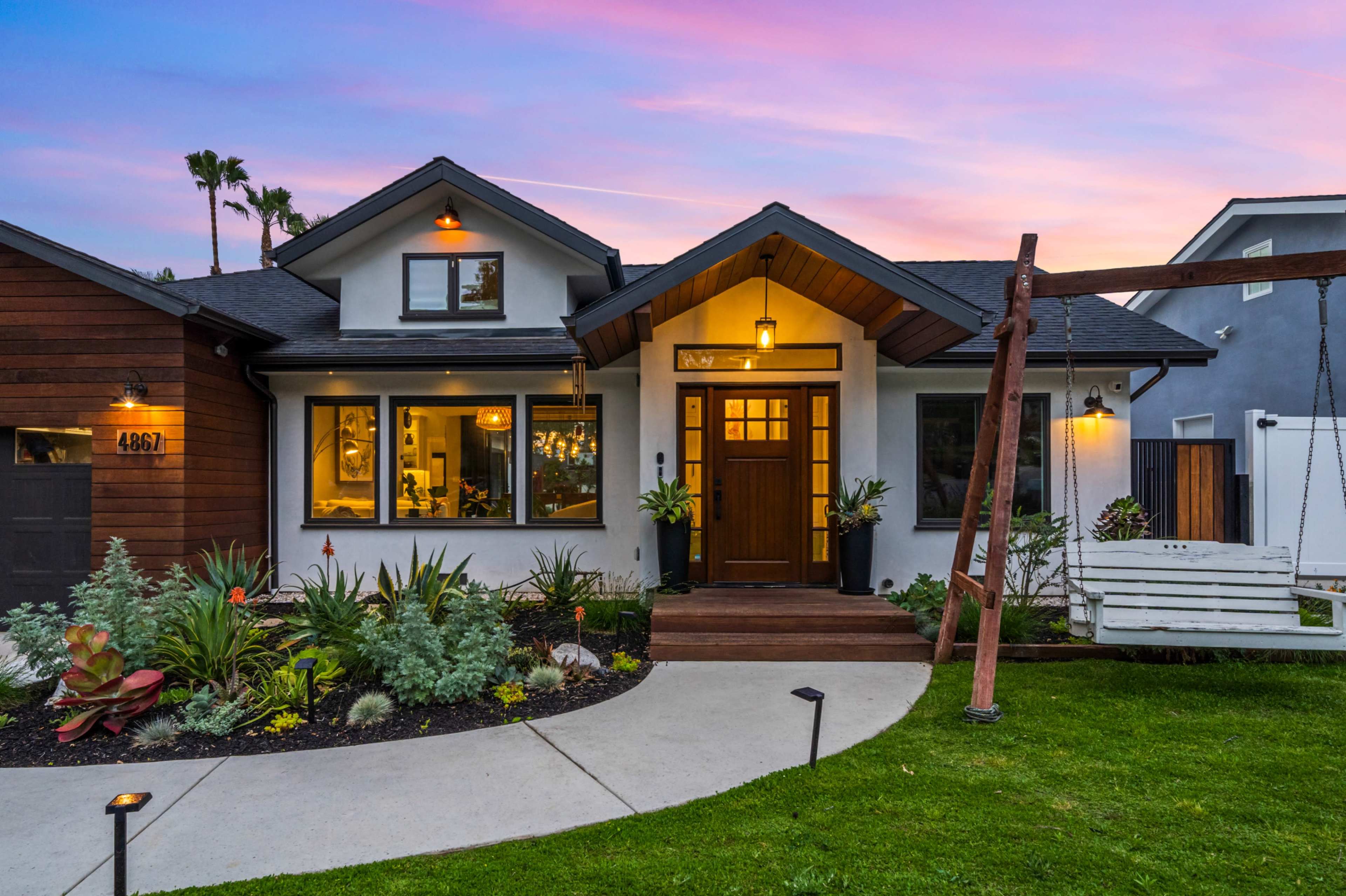 A contemporary home with a wooden front door and a swing hangs in the front yard, surrounded by landscaped greenery and a pathway leading to the entrance.
