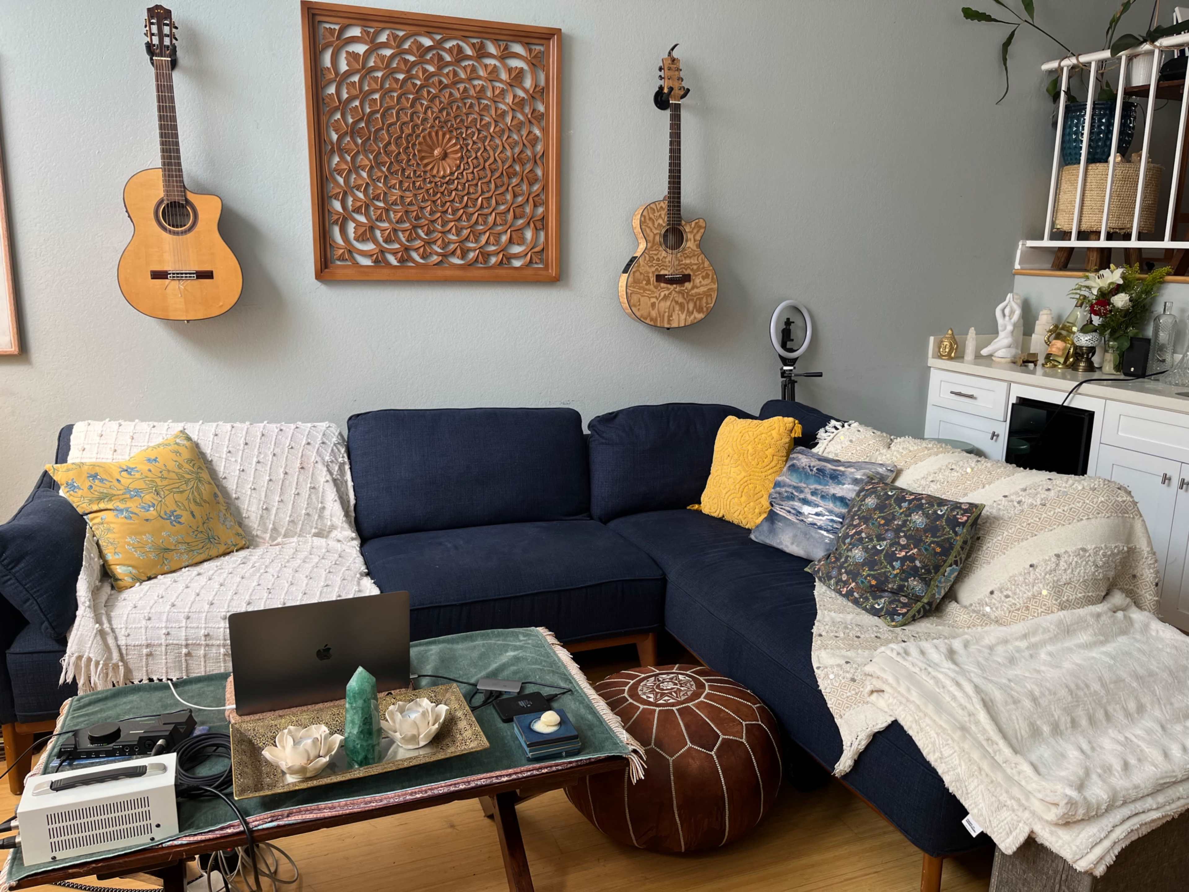 A living room with a dark blue sectional sofa, two guitars hanging on the wall, a coffee table with various items, and a decorative wall feature.