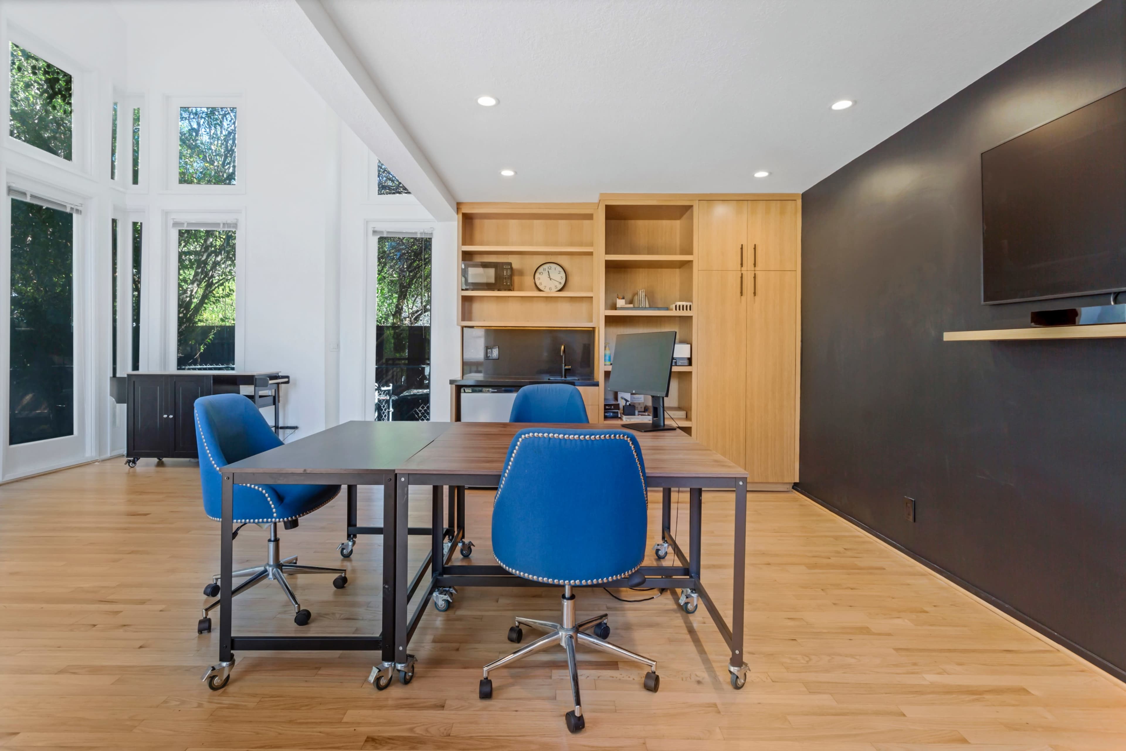 The image shows a modern workspace with four blue chairs around two tables, a wooden bookshelf, and large windows allowing natural light.