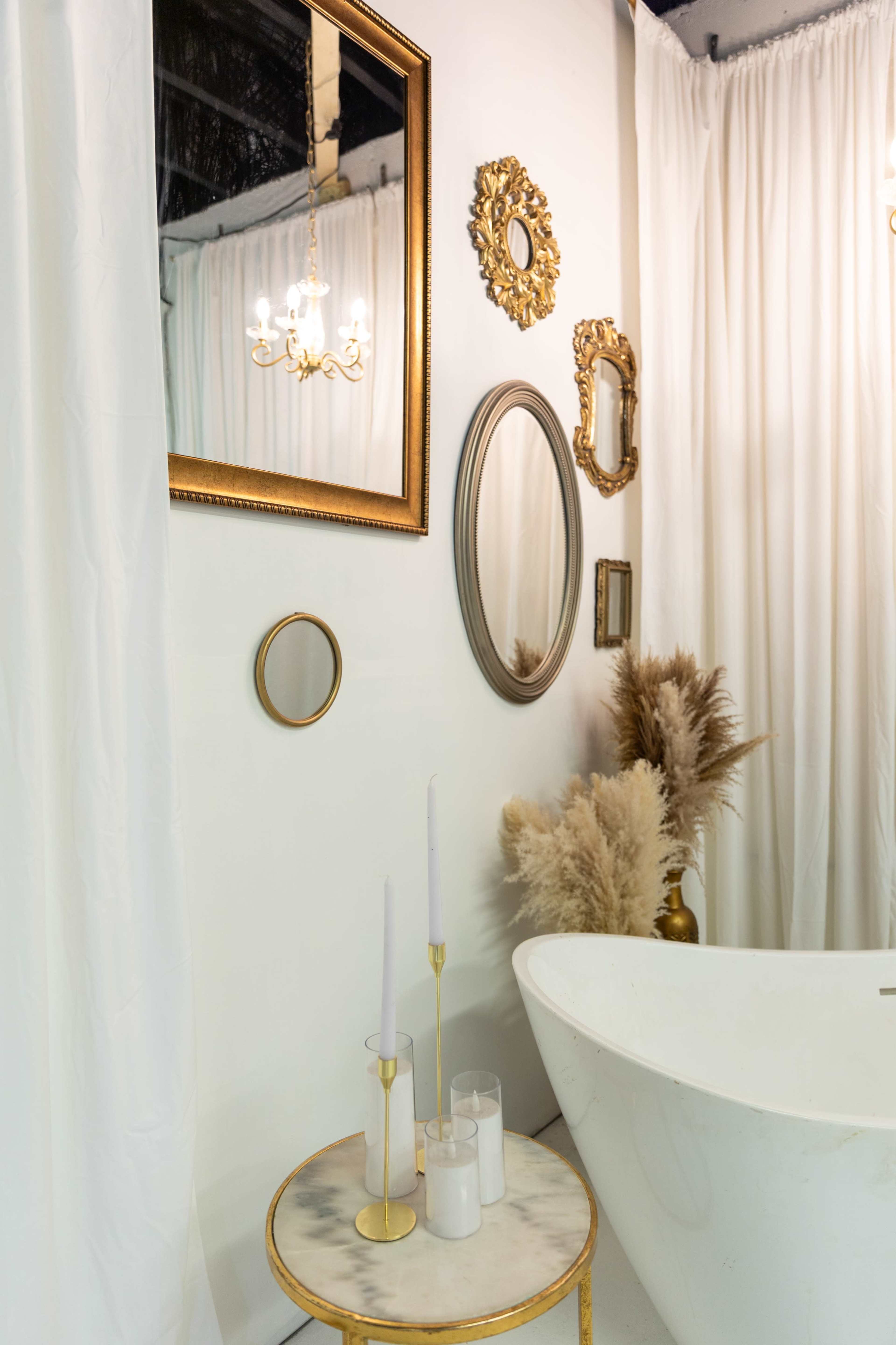 The image features a modern bathroom with a freestanding bathtub, various ornate mirrors, and decorative elements, including a small table with candles and a bouquet of dried plants.