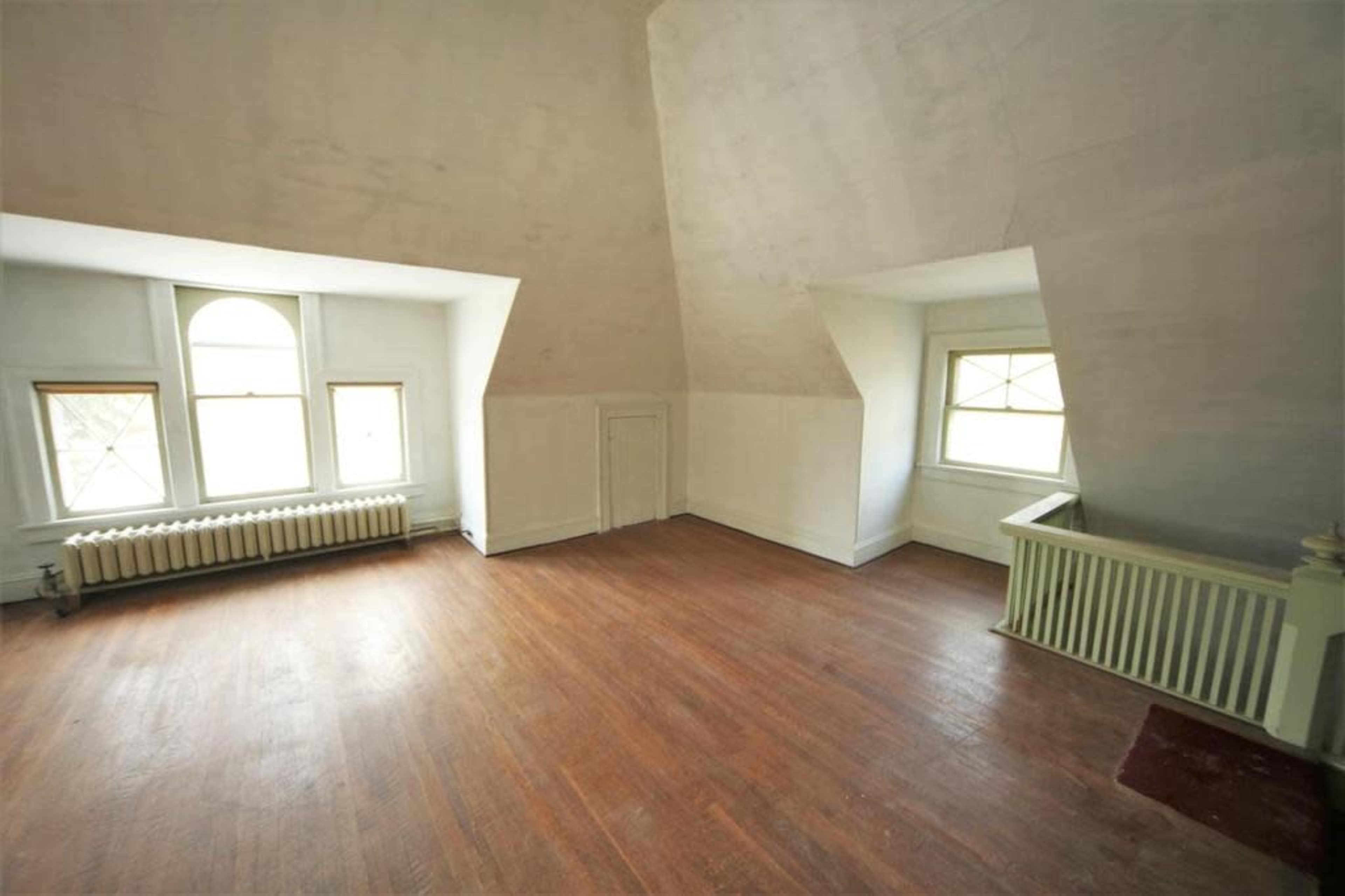 A spacious, empty room with sloped walls, wooden floors, and two windows allowing light to enter.