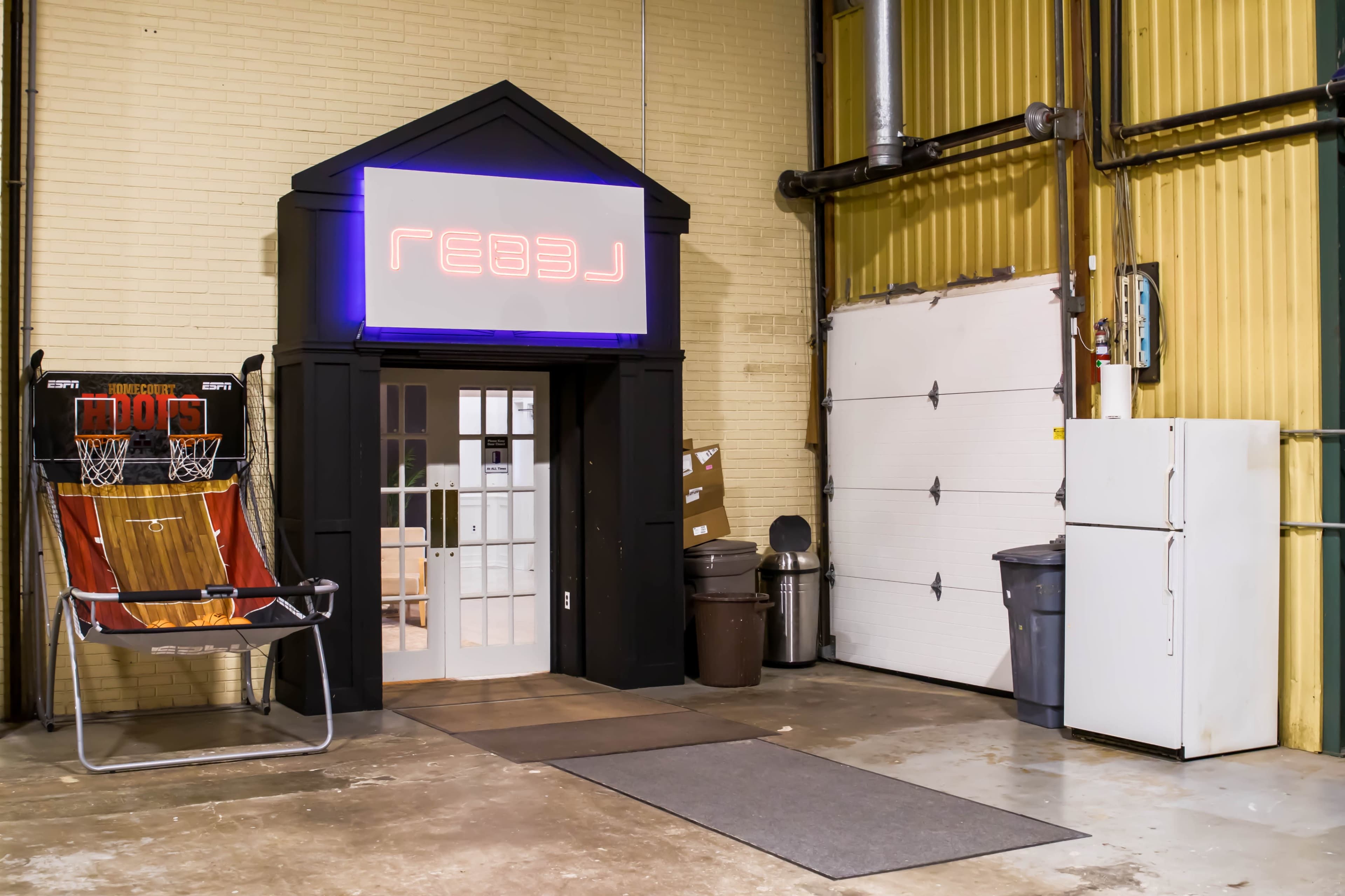 An entrance with a neon sign reading "REBEL," next to a basketball game machine and a refrigerator in a warehouse-like space.