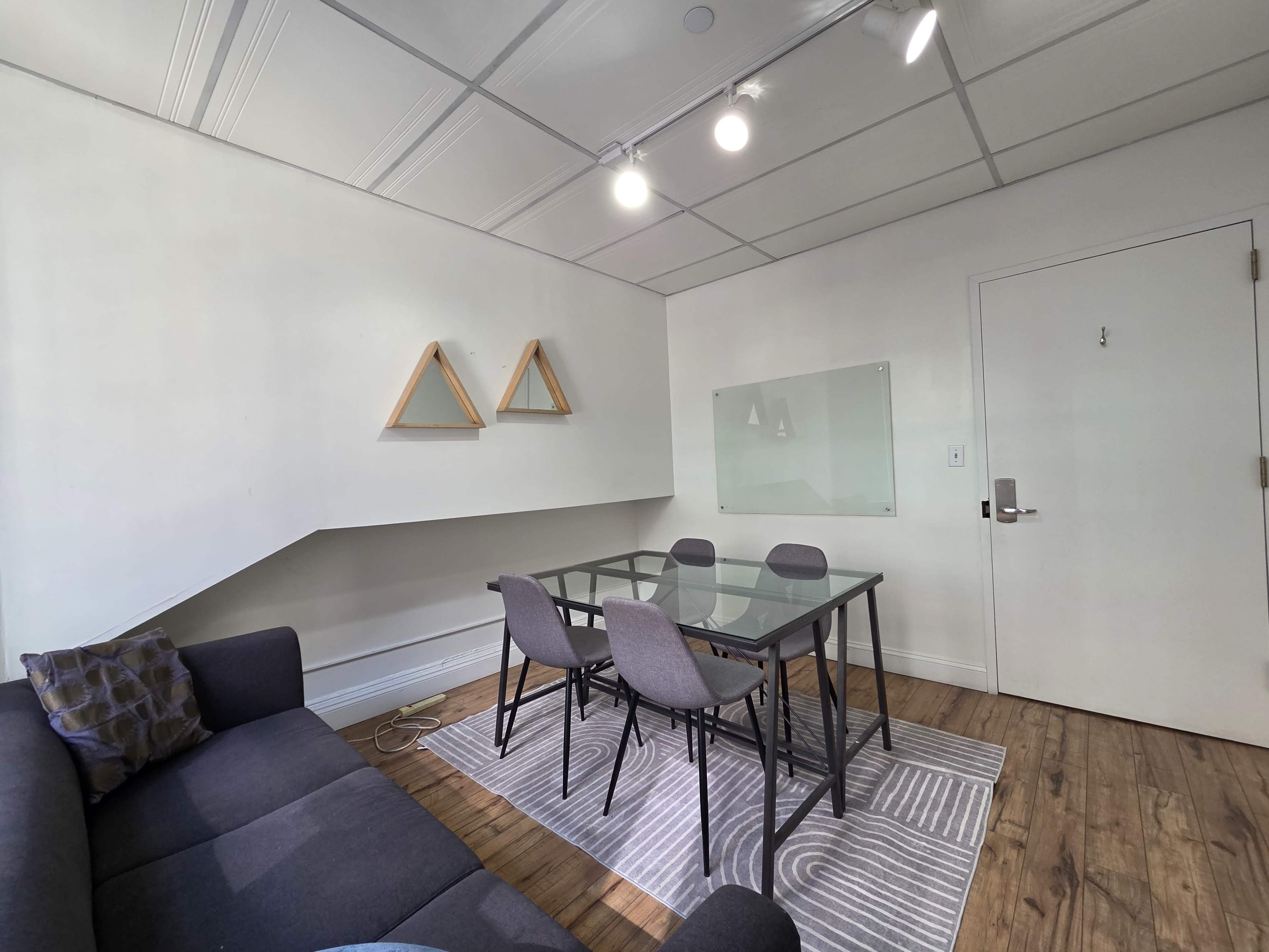 The image shows a small, modern room featuring a glass dining table with four chairs, a sofa, a rug, and decorative wall shelves.