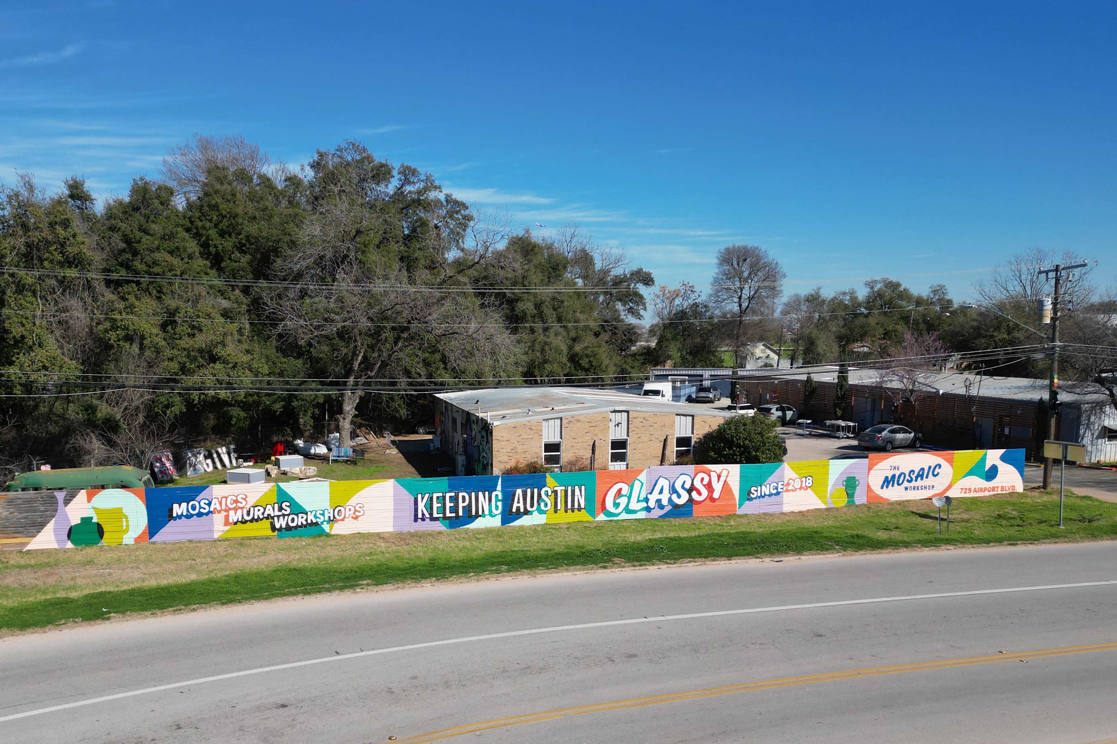 A brightly colored mural on a fence promotes "Mosaic Murals Workshops" in Austin, Texas, alongside the slogan "Keeping Austin Glassy Since 2018" against a clear blue sky.