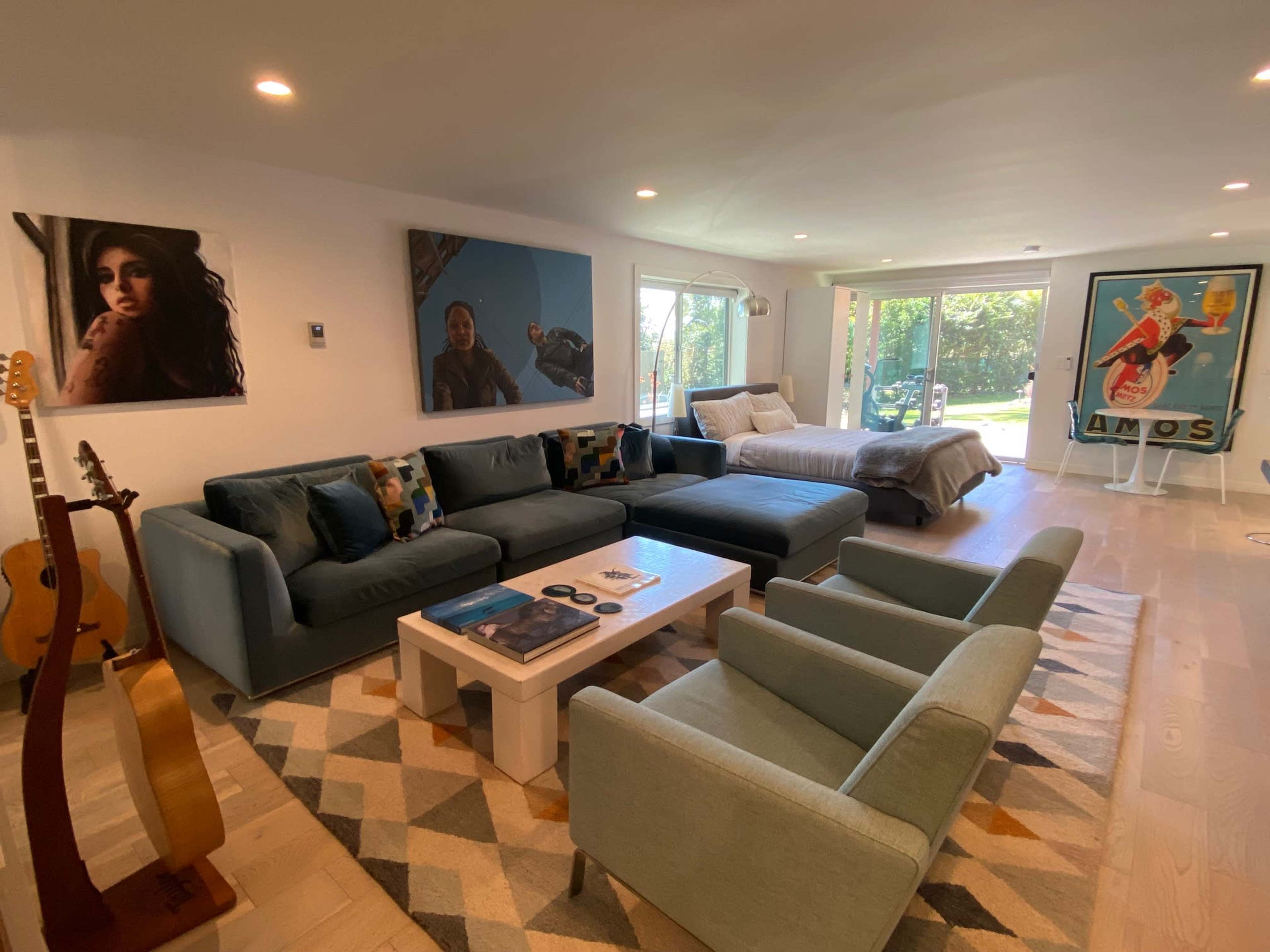 A spacious living room features a sectional sofa, decorative pillows, a coffee table, artwork on the walls, and large windows that open to a garden view.