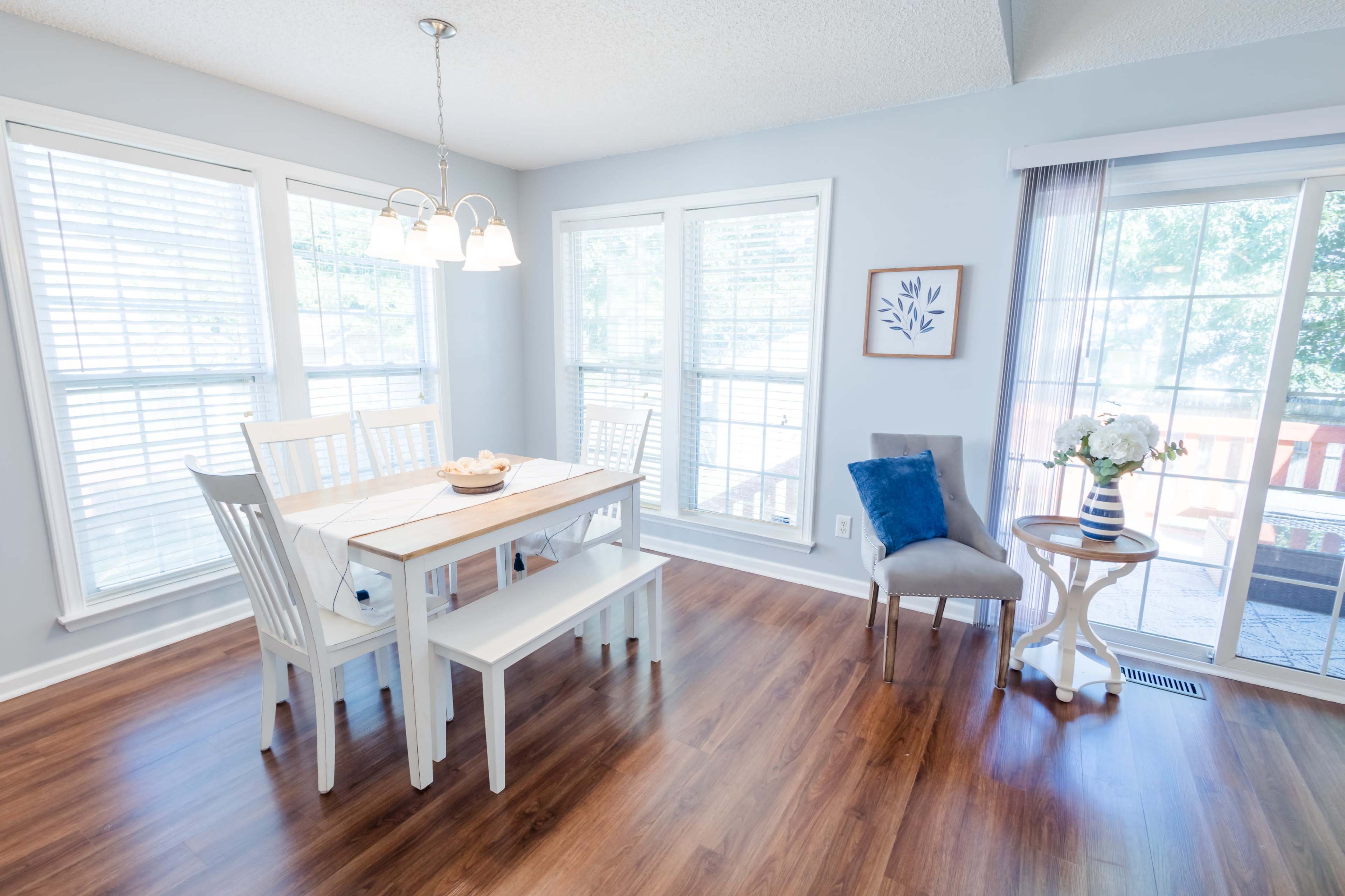 A dining area features a white table with chairs and a bench, surrounded by large windows letting in natural light.