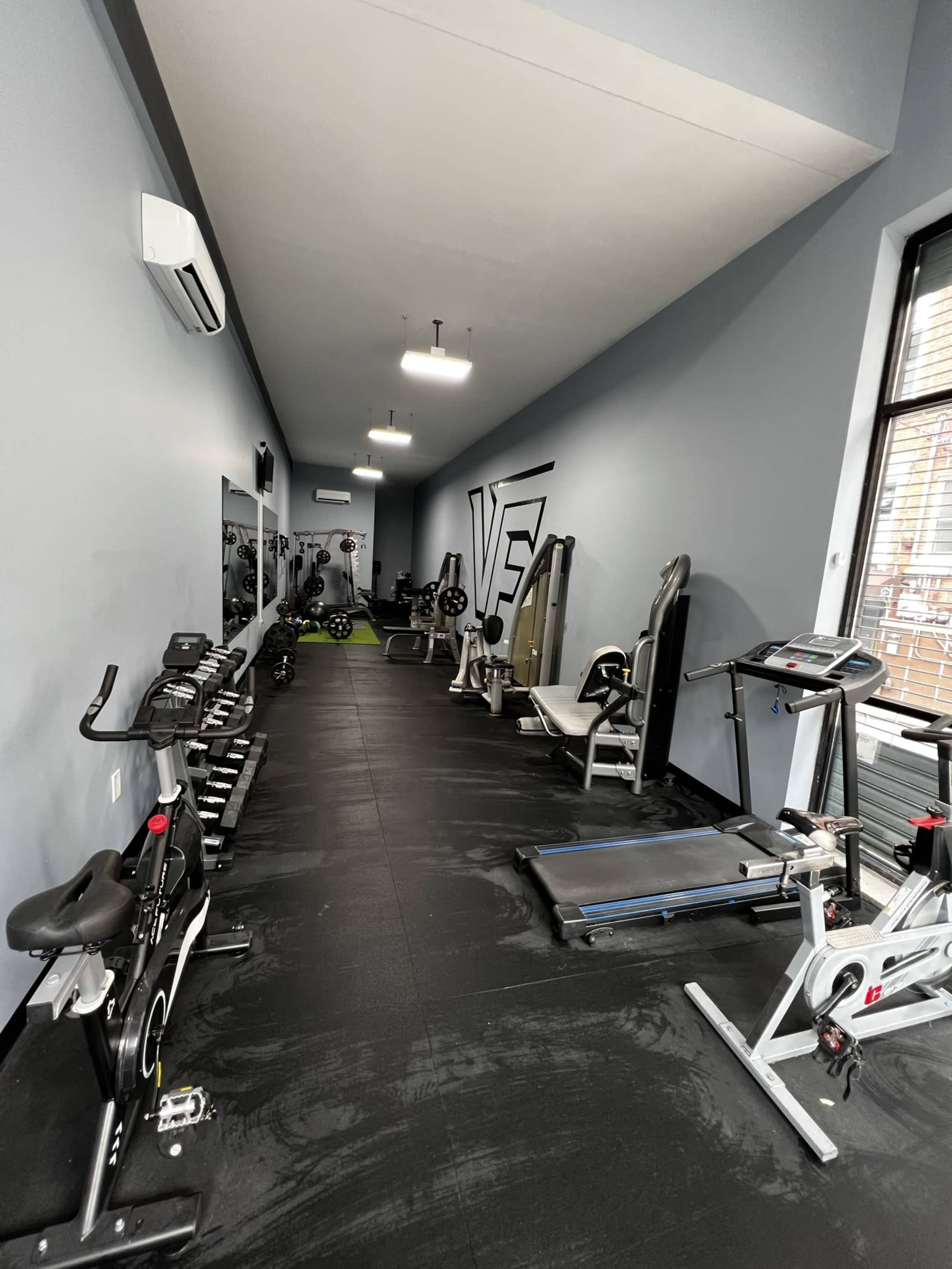 The image shows a narrow gym space equipped with various exercise machines and weights along the walls.
