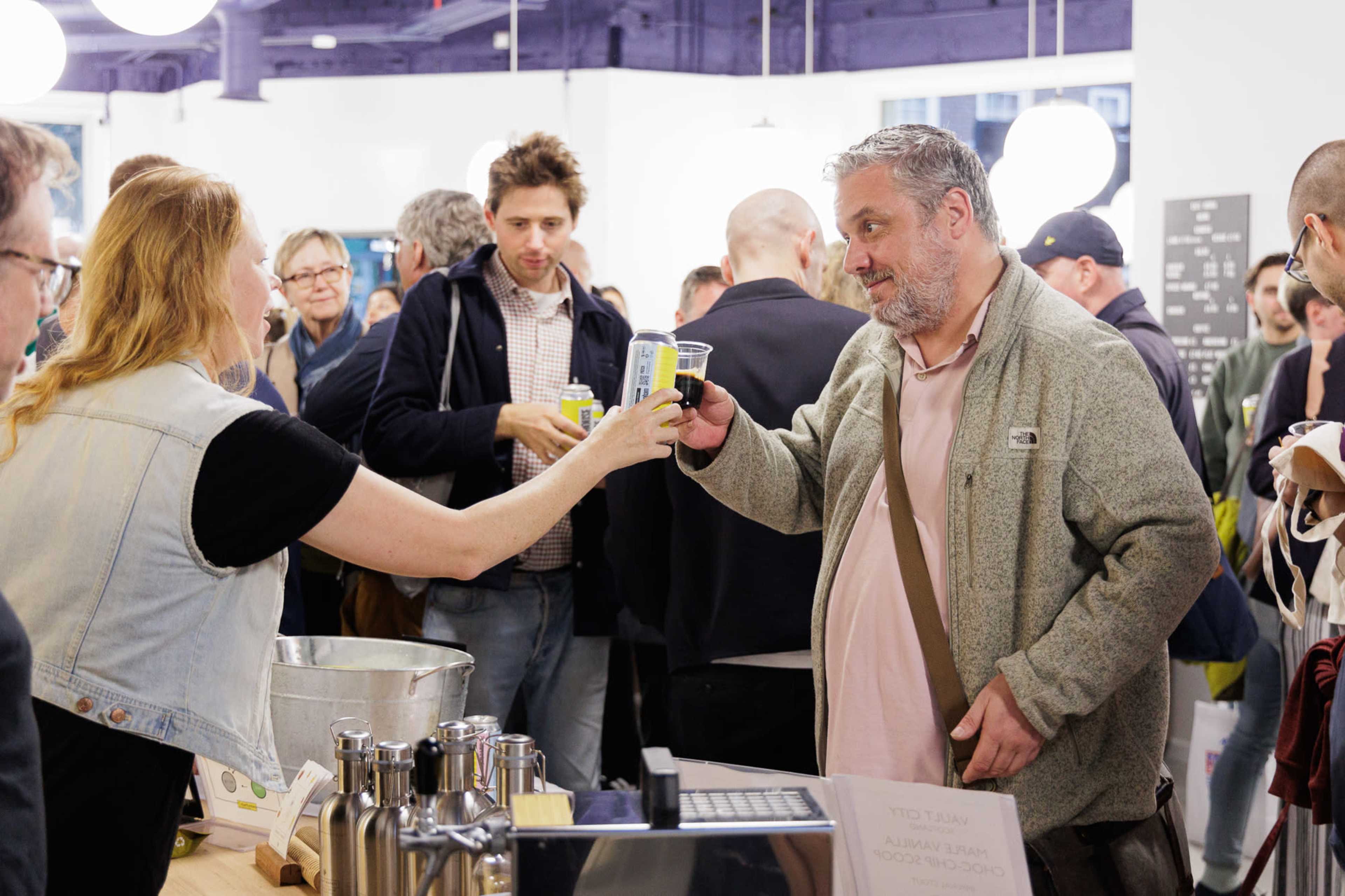 A person hands a beverage to another individual at an indoor event with people mingling in the background.