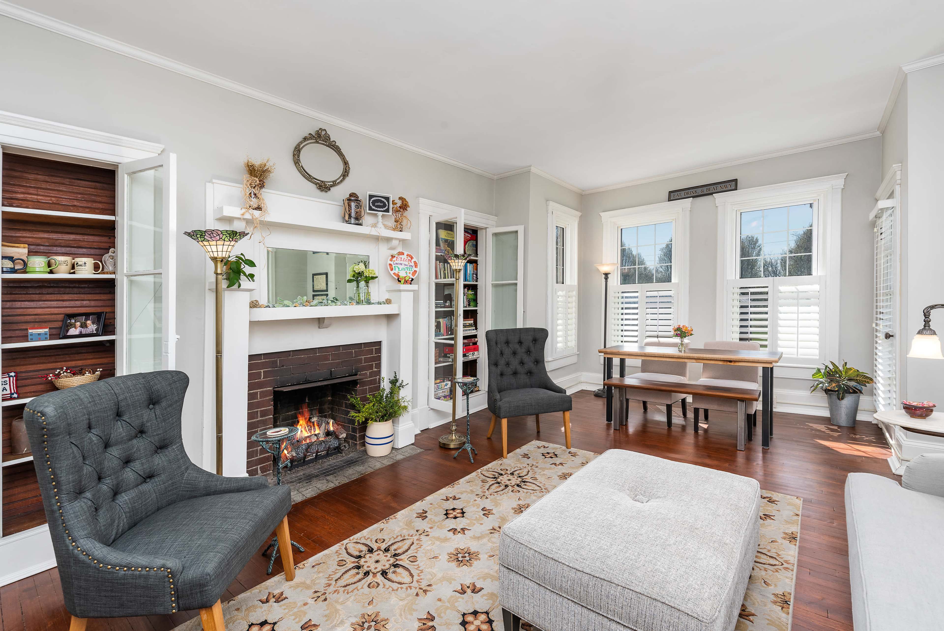 A cozy living room features a fireplace, two upholstered chairs, a coffee table, and a dining area with natural light coming through the windows.