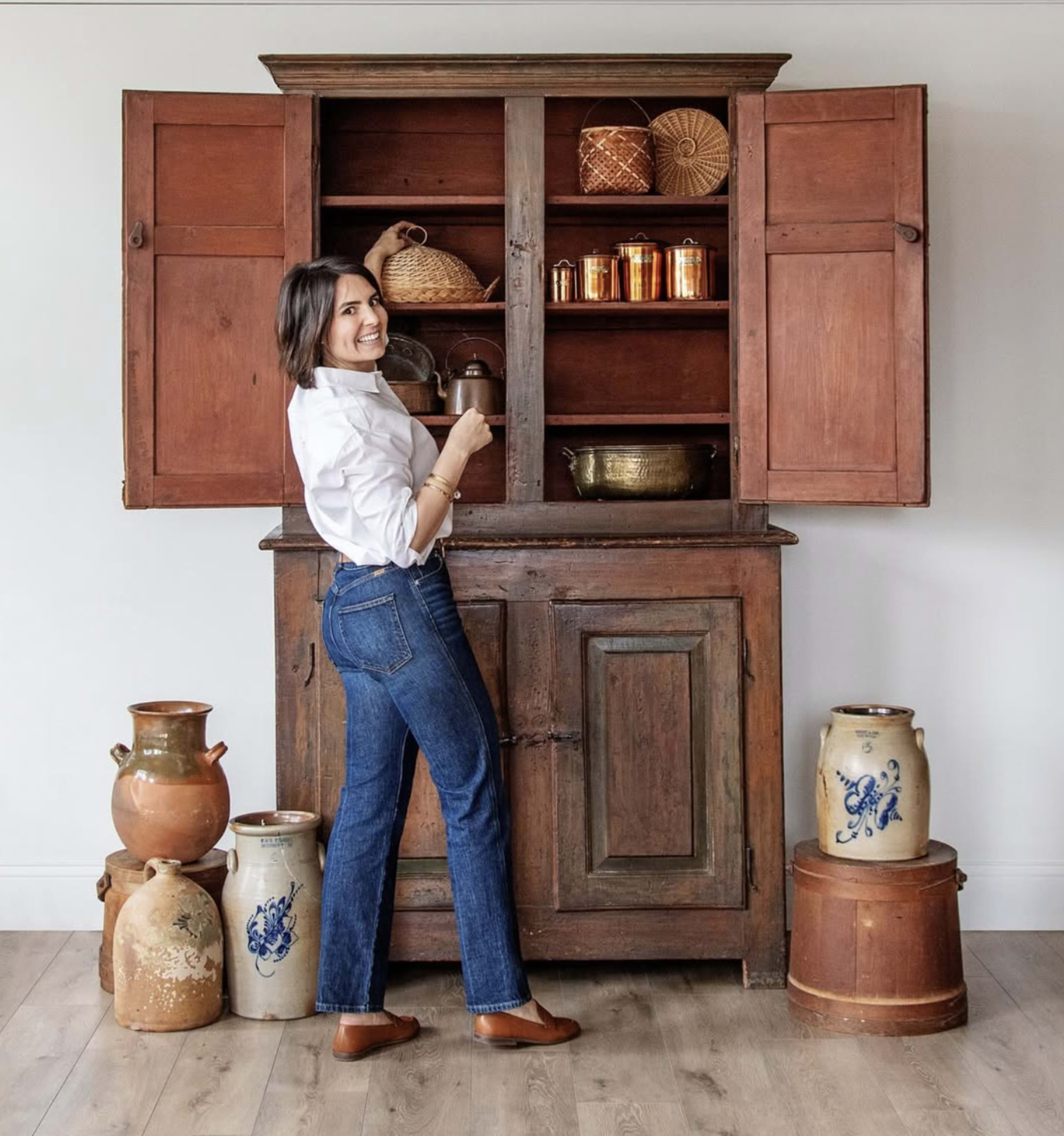 A woman stands beside a vintage wooden cabinet filled with various decorative items, surrounded by ceramic containers on the floor.