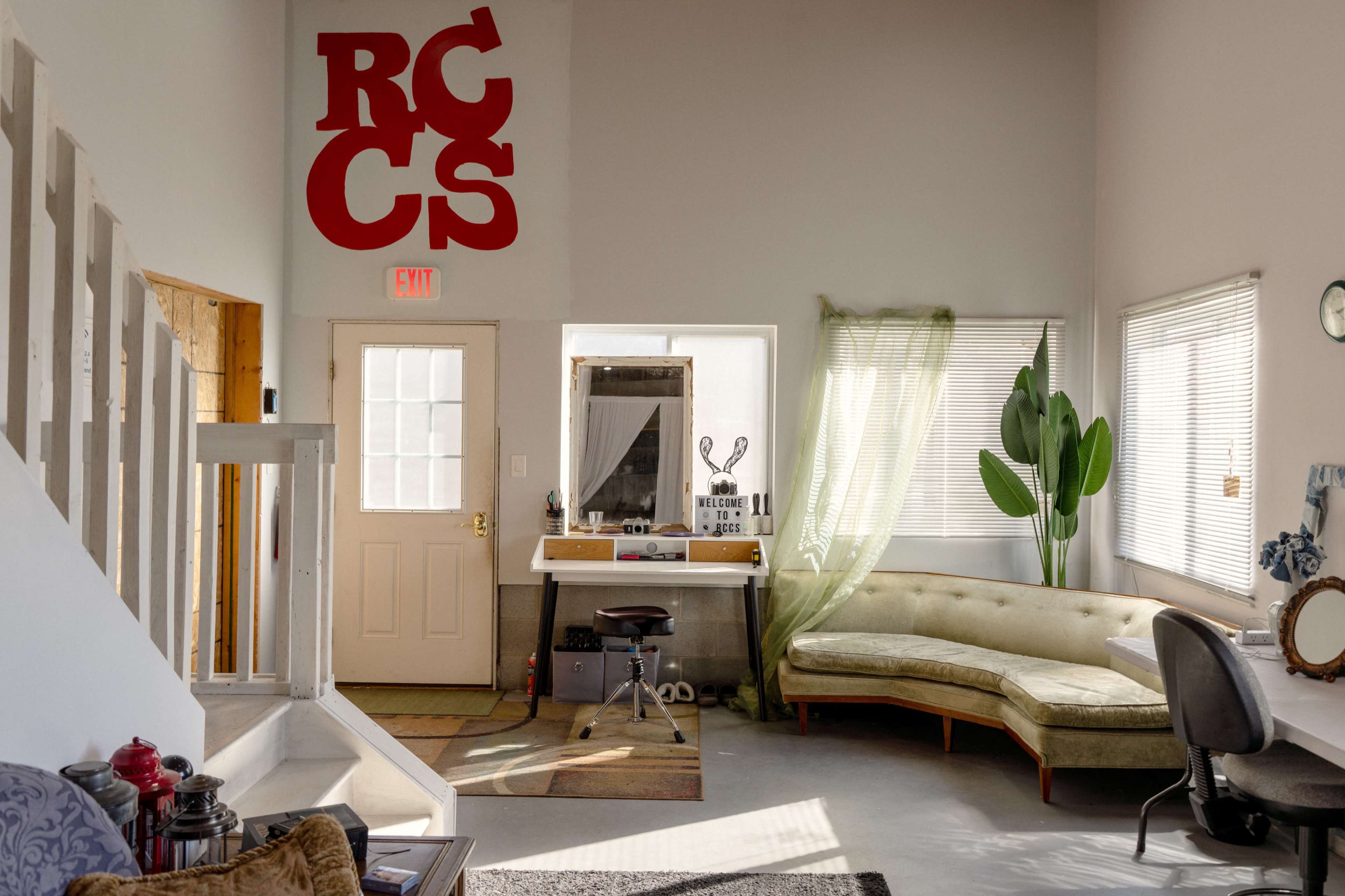 The image shows a bright interior space featuring a green couch, a workspace with a desk, and decorative plants, all under a high ceiling with a red letter sign against the wall.