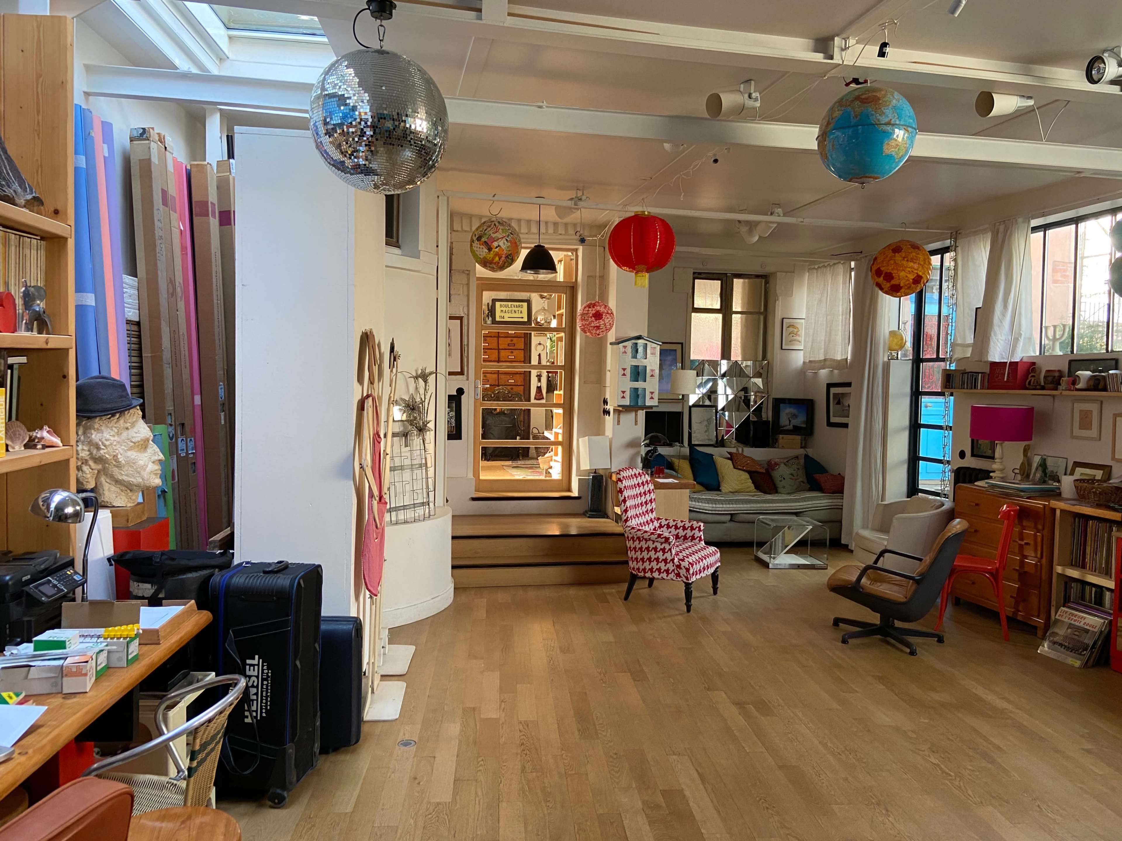 The image shows a spacious, well-lit studio or creative space with various decorative elements, seating areas, and a staircase leading to a door.
