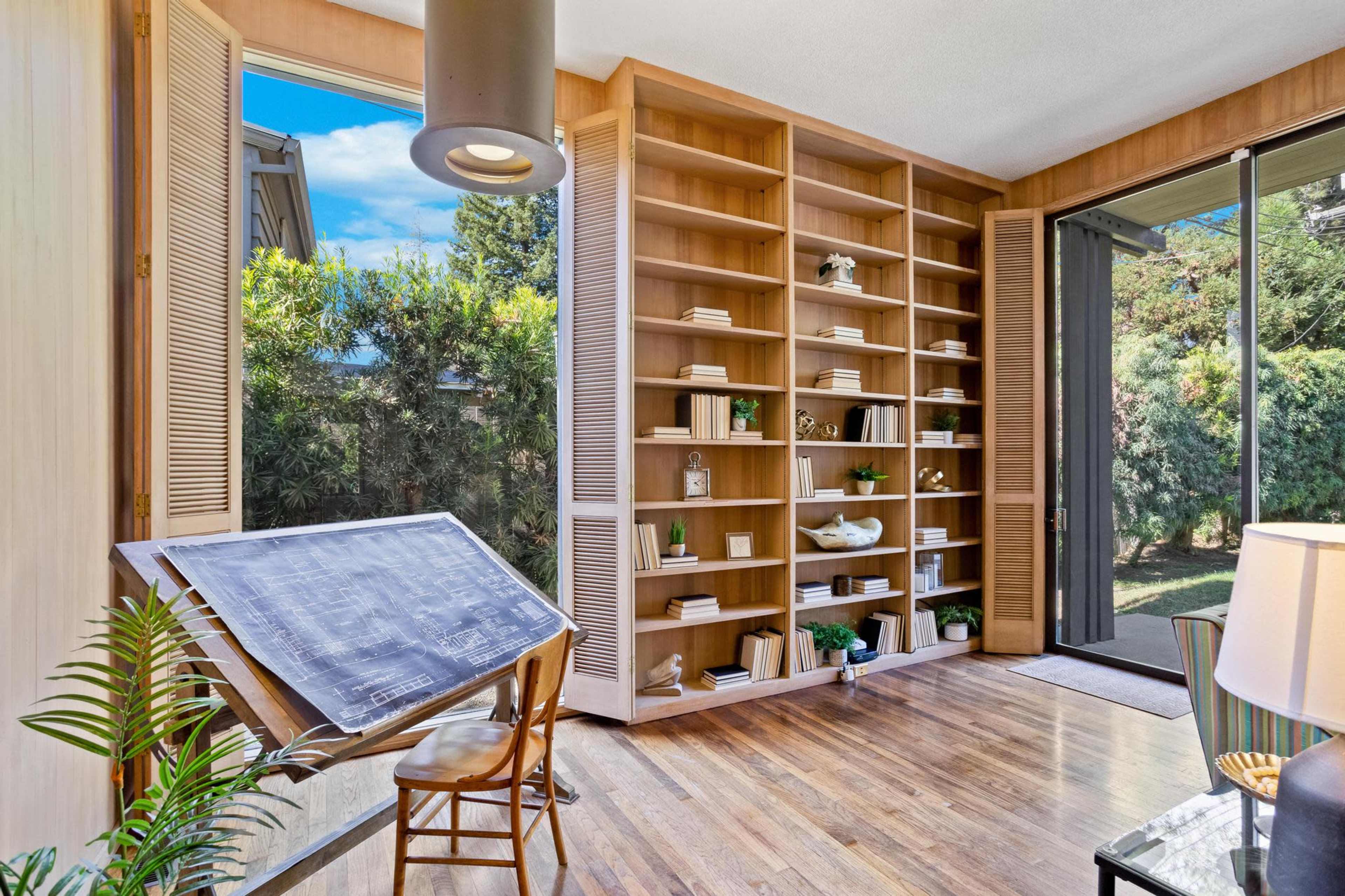A bright room featuring a wooden bookshelf filled with various items, a drawing table in the foreground, and large windows overlooking a green outdoor space.