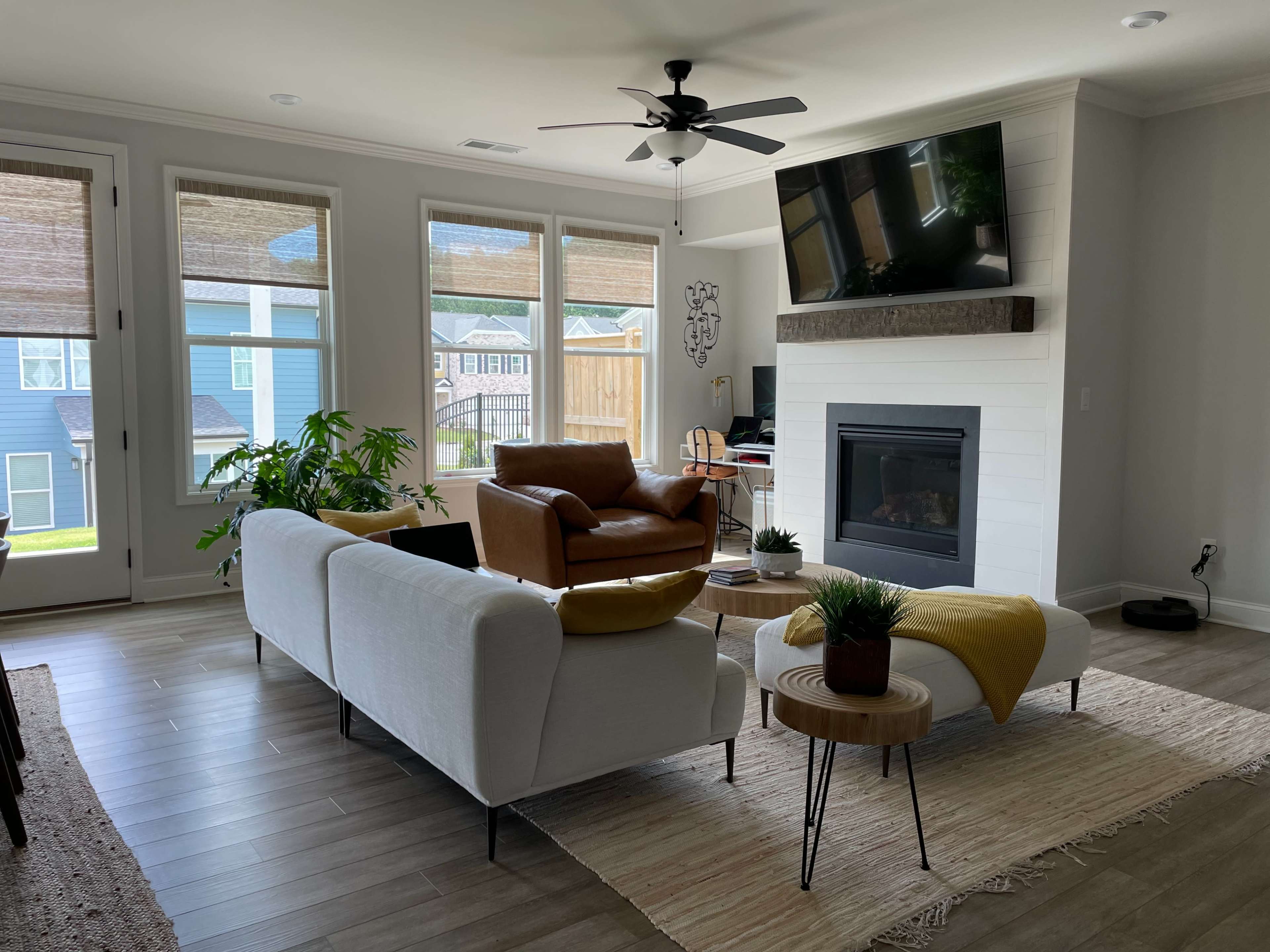 The image shows a bright living room with a white sofa, a brown leather armchair, a fireplace, and large windows overlooking a green yard.