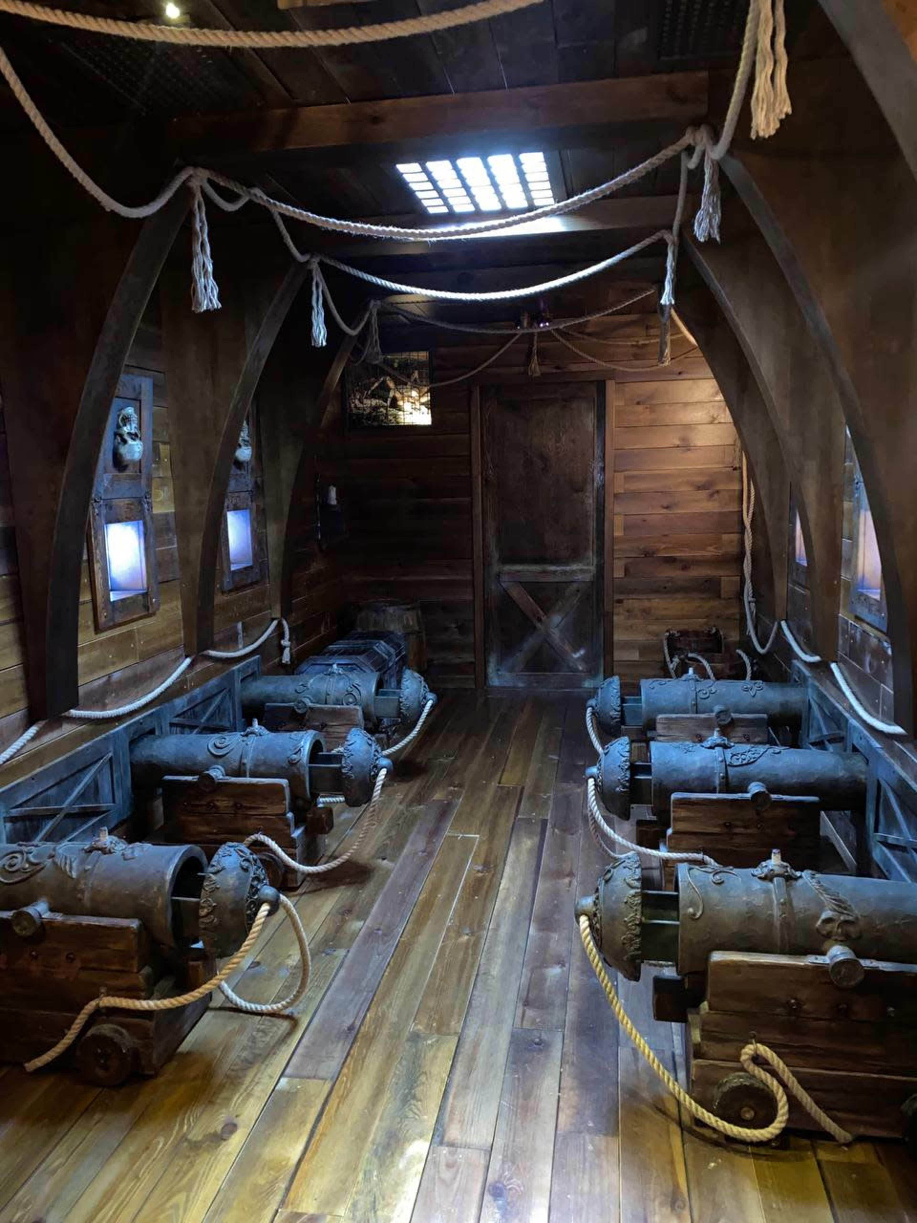 The interior of a ship features several cannons arranged on a wooden floor, with wooden walls and ropes hanging from the ceiling.