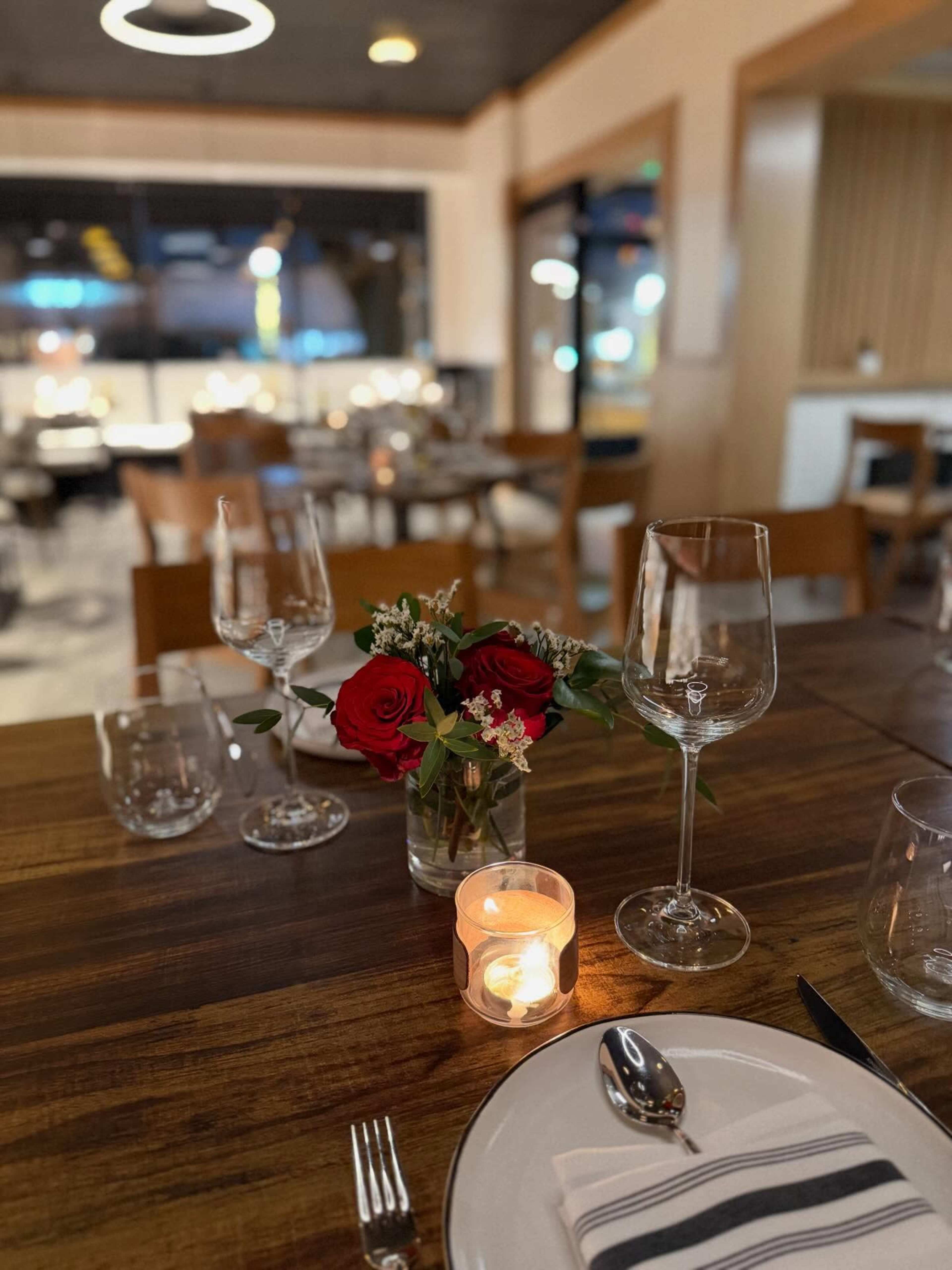 A dining table is set with glassware, a small vase of red roses, and a lit candle, all arranged in a warmly lit restaurant.