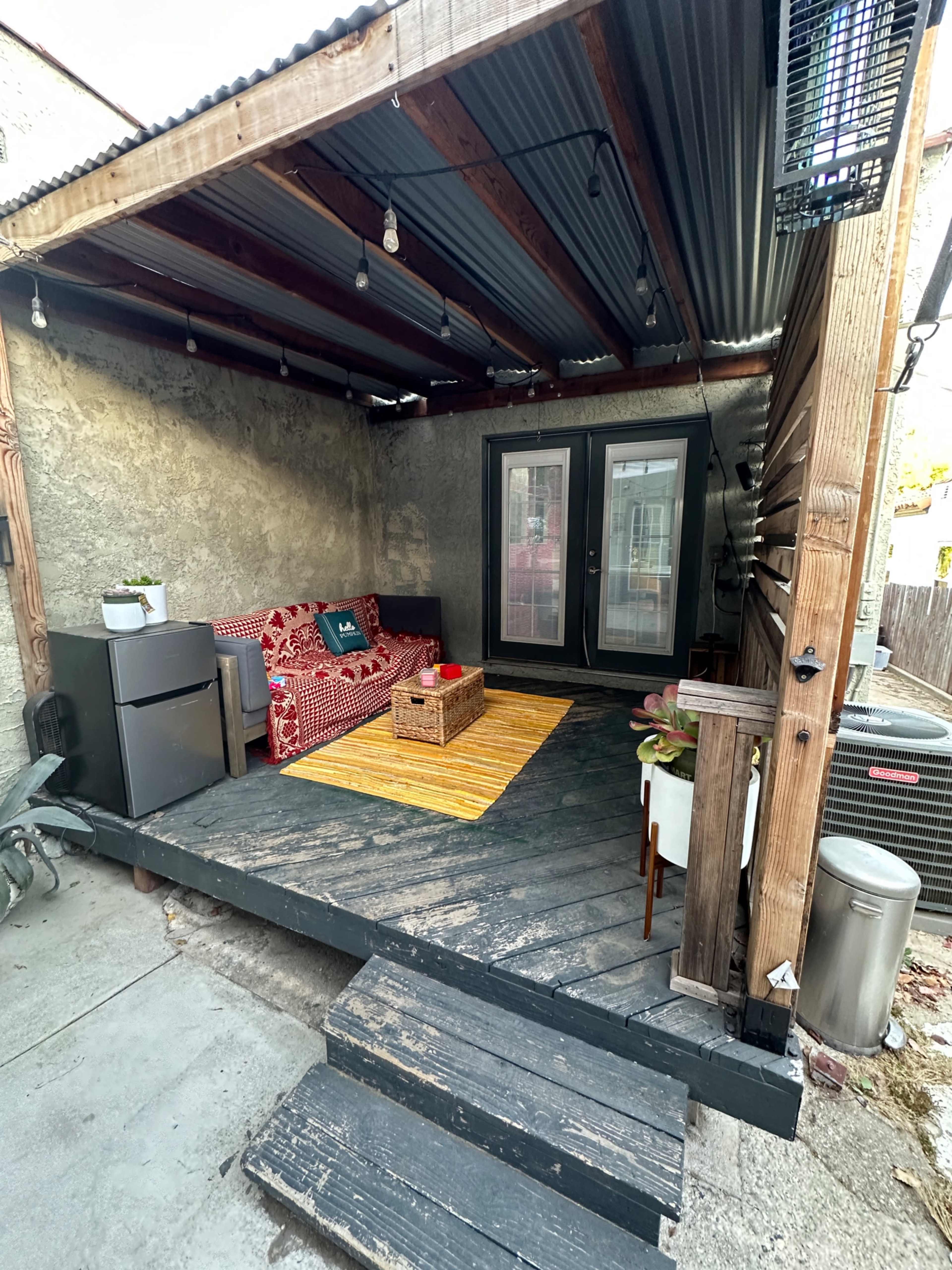 A covered patio with a wooden deck, featuring a sofa with red cushions, a coffee table, a small refrigerator, and potted plants.