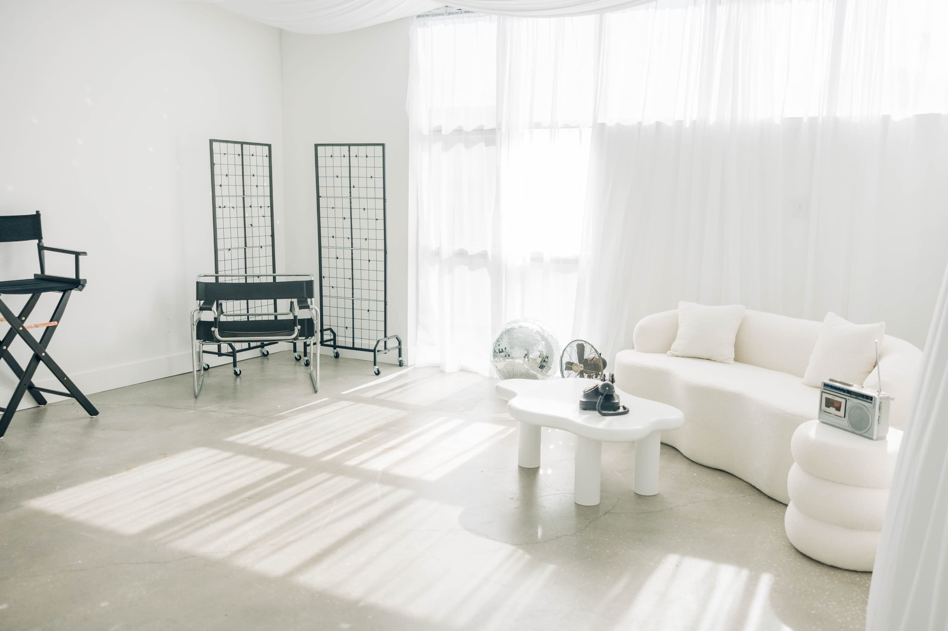 The image shows a modern, minimalist room with a white sofa, a coffee table, a director's chair, and light filtering through sheer curtains.
