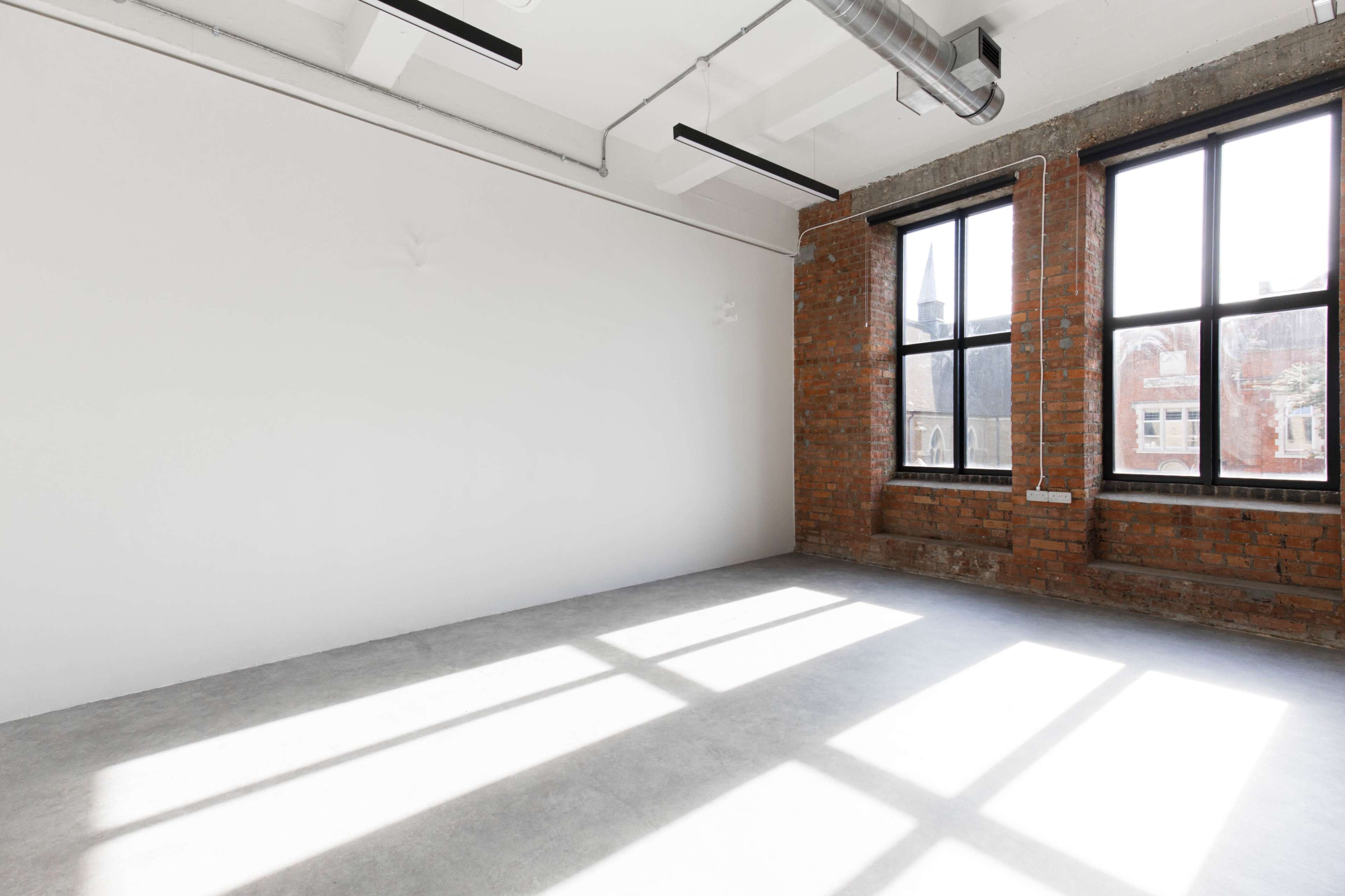 The image shows an empty interior space with a concrete floor and large windows allowing natural light to cast shadows on the walls.
