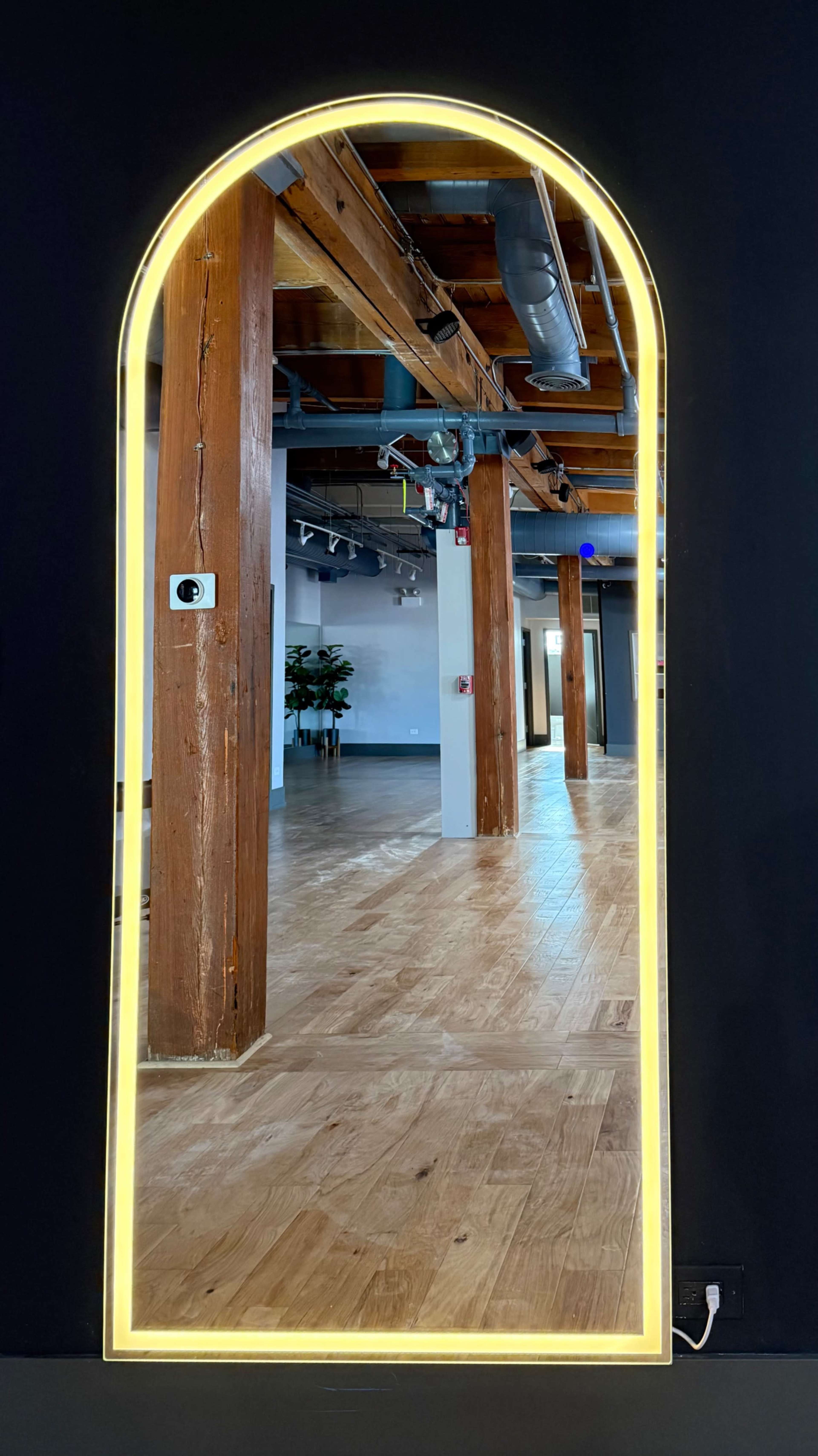A tall, arched mirror with illuminated edges reflects a spacious interior featuring wooden beams and a polished floor.