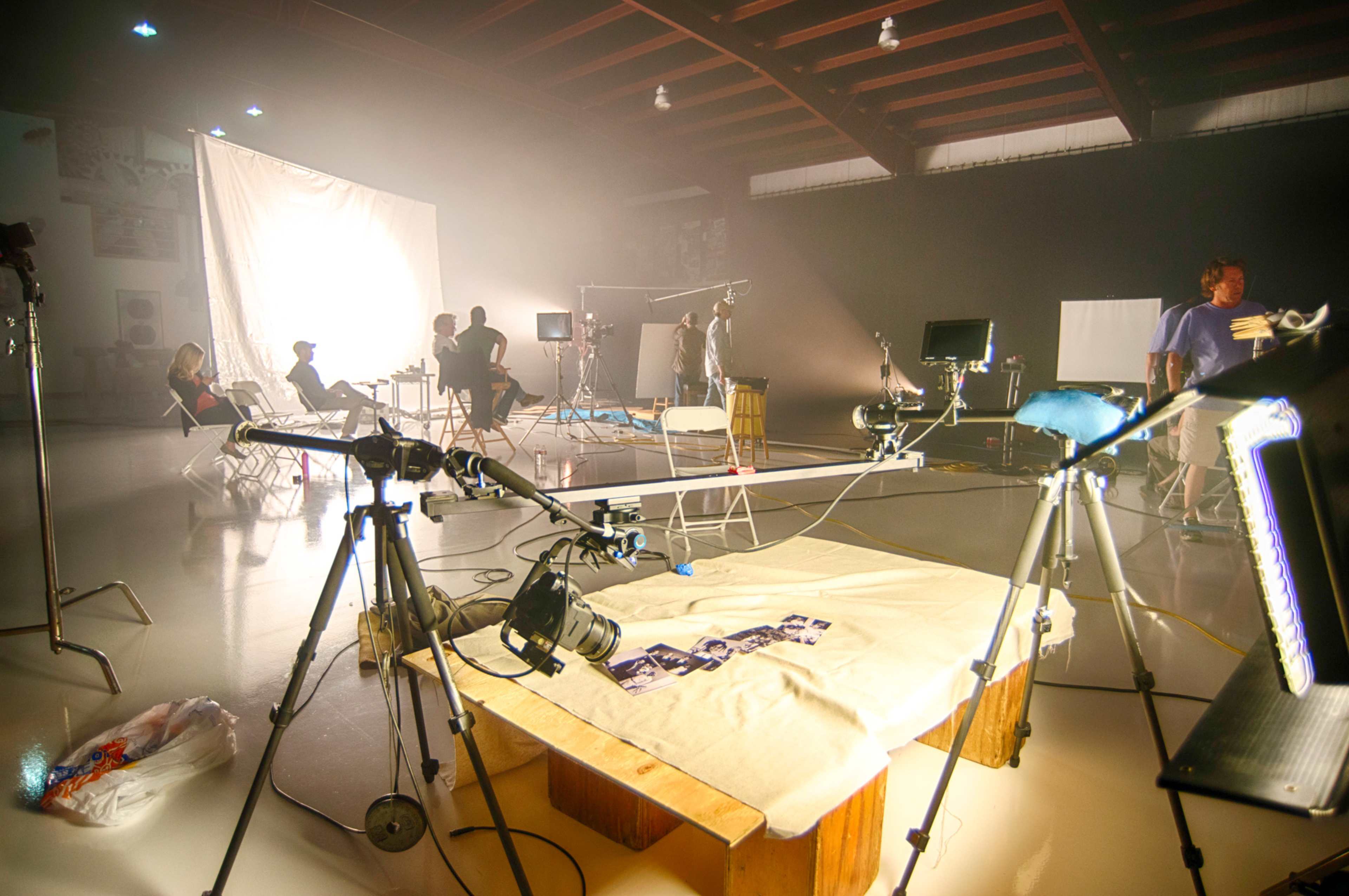 A photography studio setup features several camera tripods, lighting equipment, and people engaged in a photo shoot under soft, diffused lighting.