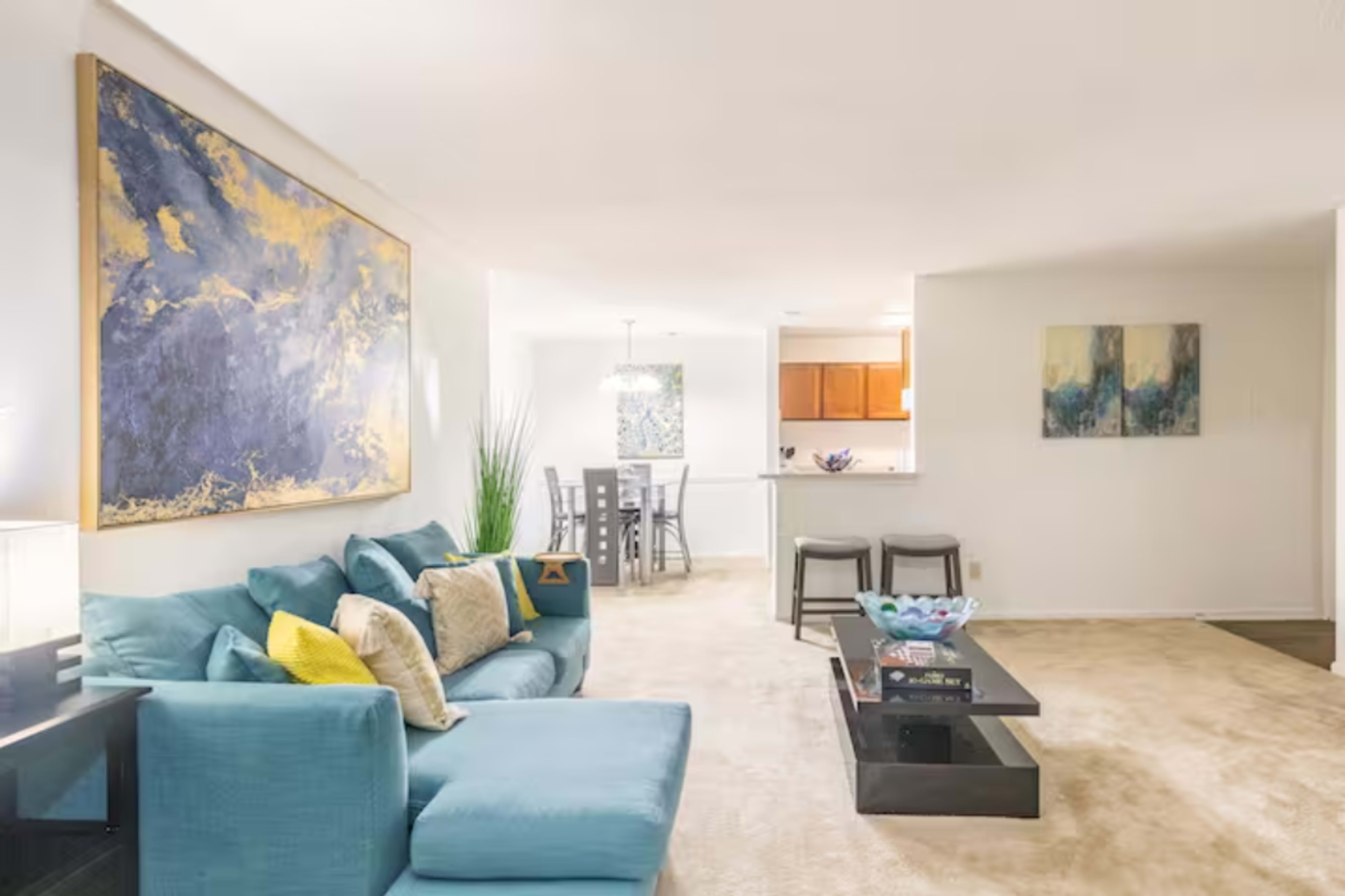A modern living room featuring a turquoise sectional sofa, a black coffee table, and a dining area visible in the background with wooden cabinets.