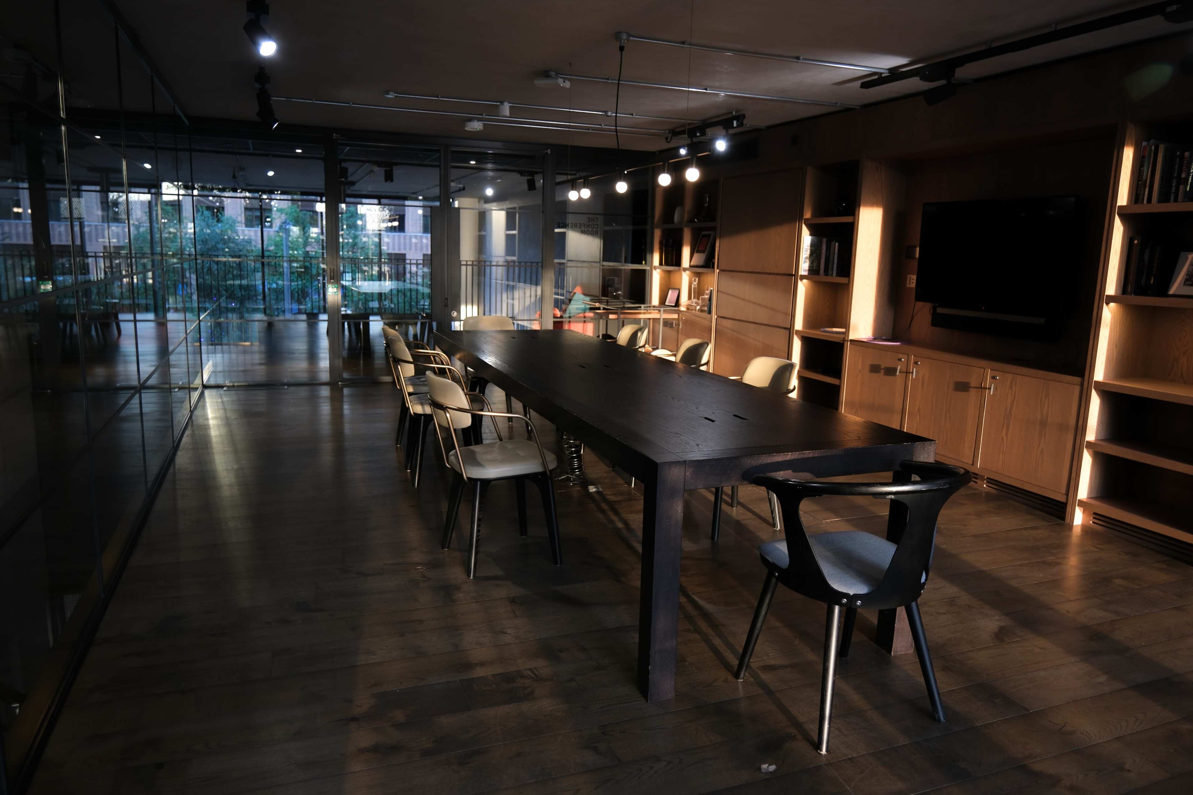A long wooden table is surrounded by chairs in a modern conference room with glass walls and shelves lined with books.