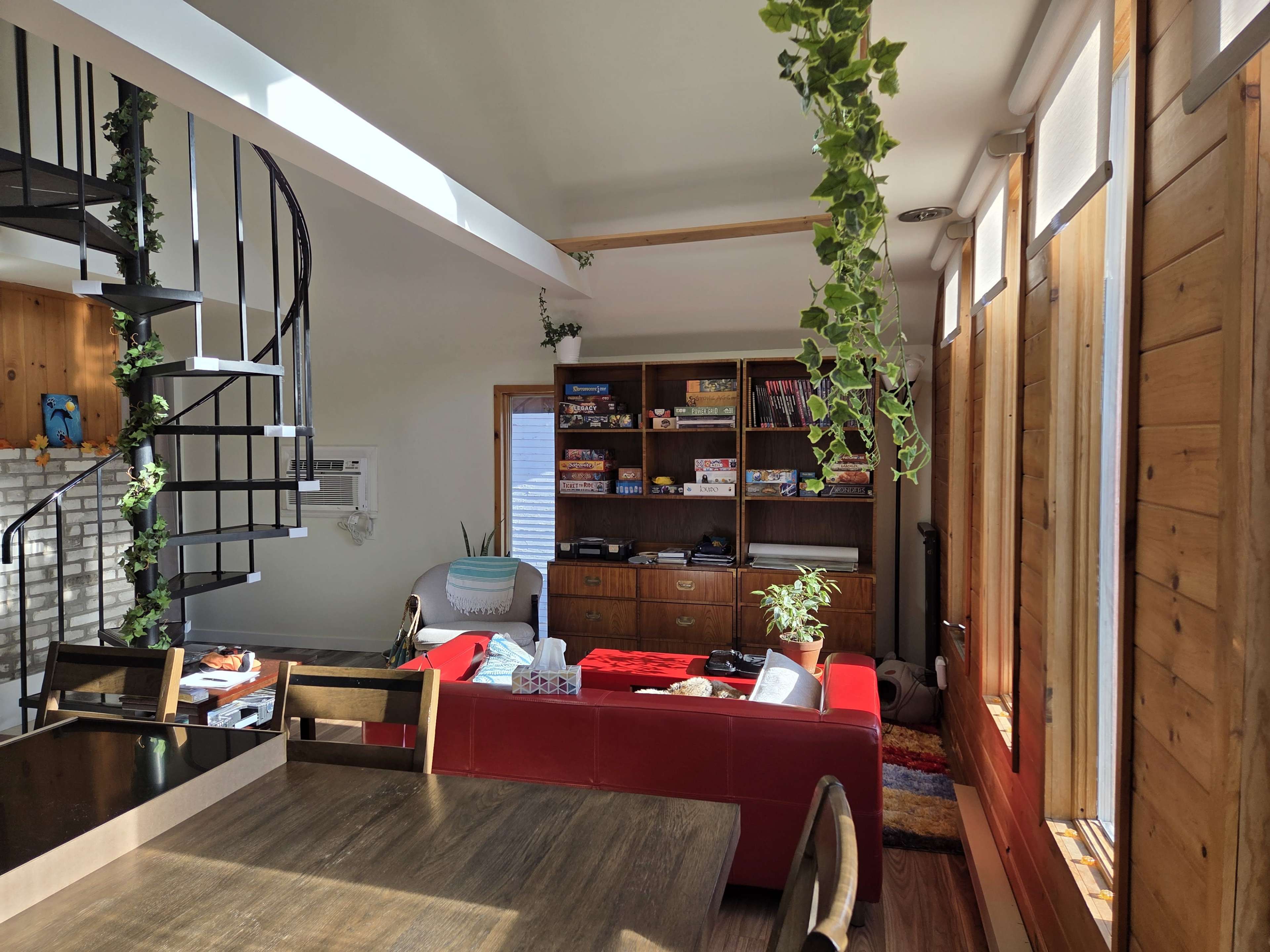 The image shows a modern interior living space featuring a red sectional sofa, a wooden dining table, a spiral staircase, and a bookshelf with games, illuminated by natural light from large windows.