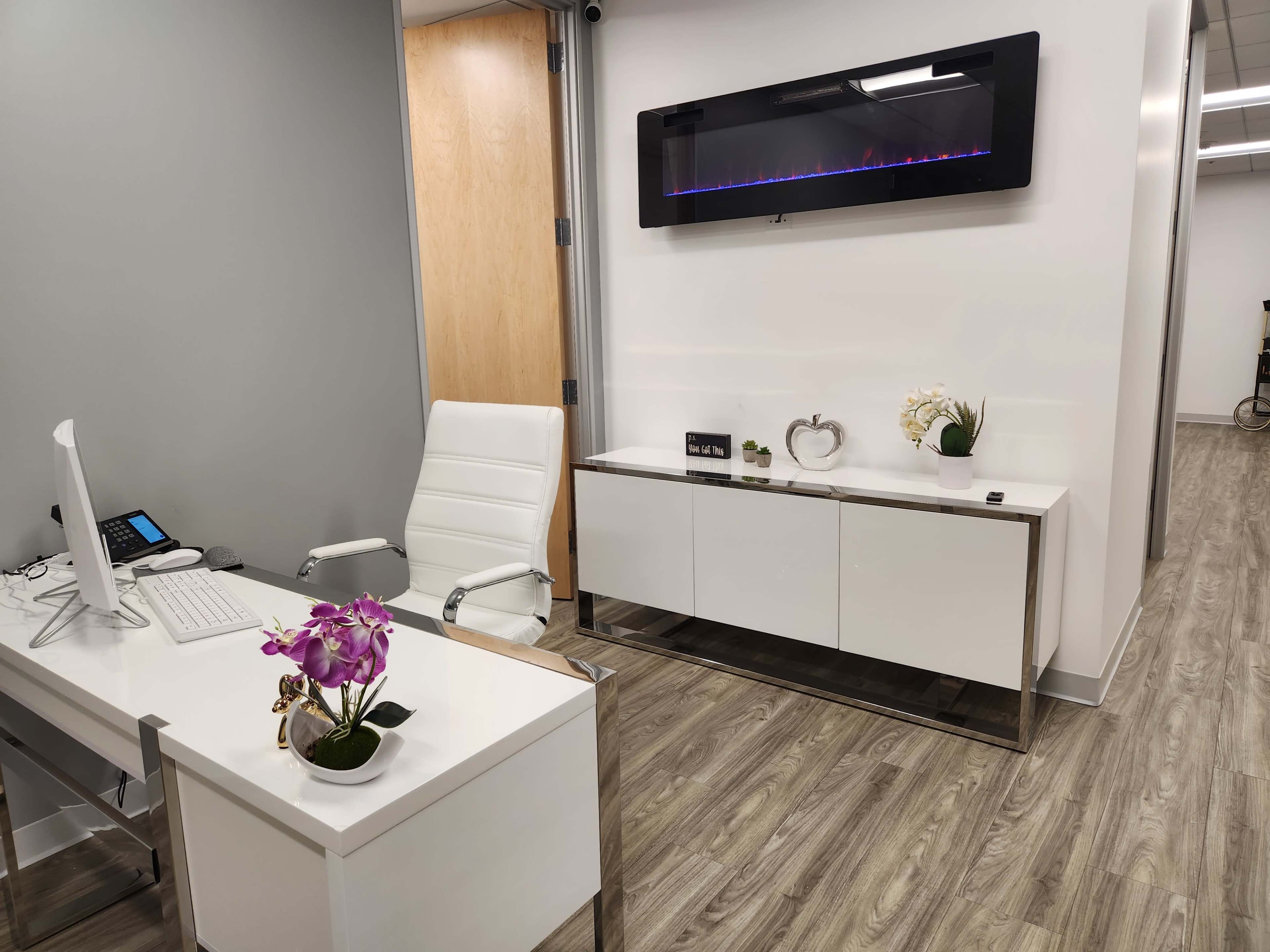A modern office space featuring a white desk with a computer, an ergonomic chair, and a sleek cabinet against a wall, complemented by decorative plants and an electric fireplace above.