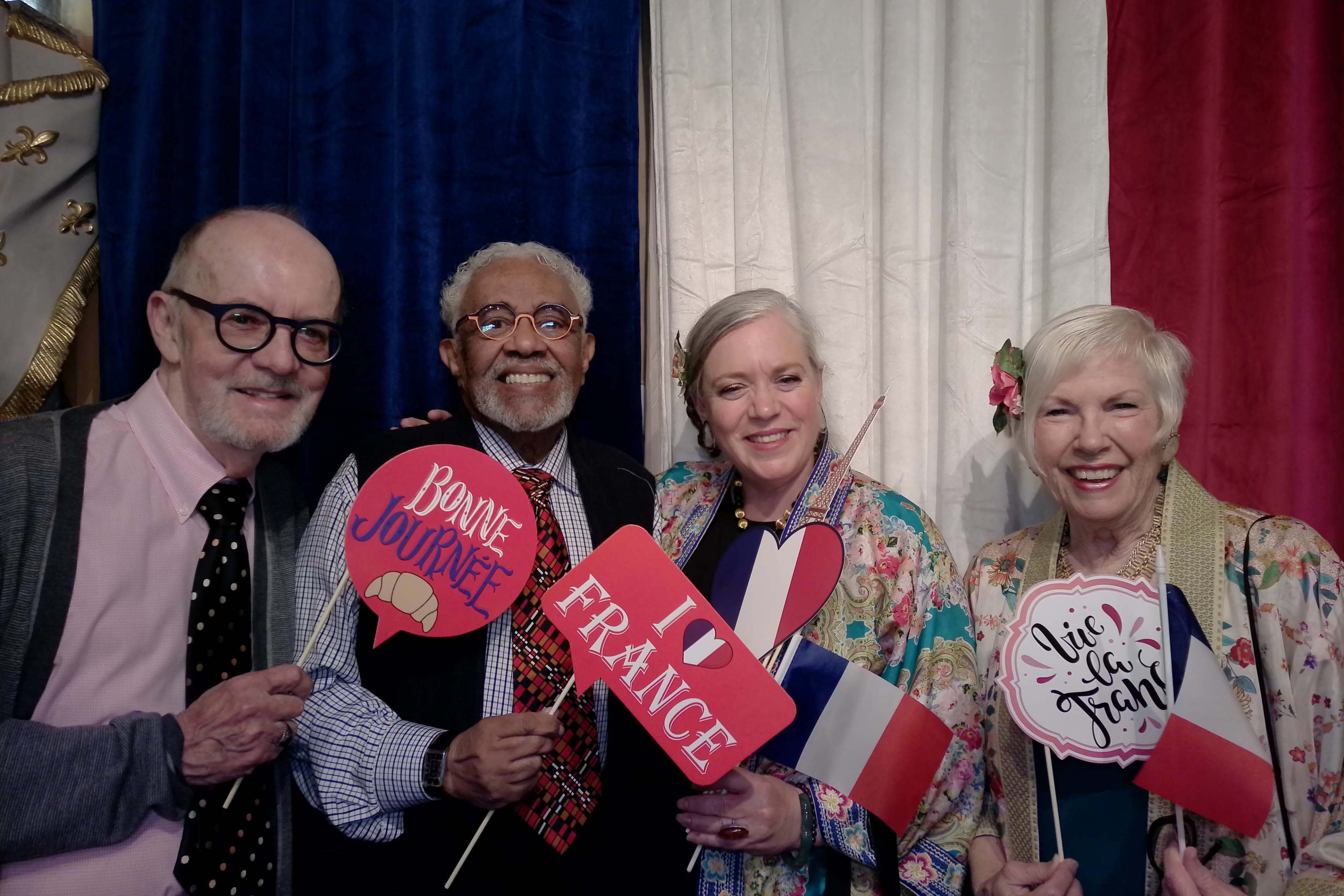 Four individuals hold decorative signs with French captions while standing in front of a backdrop featuring the colors of the French flag.
