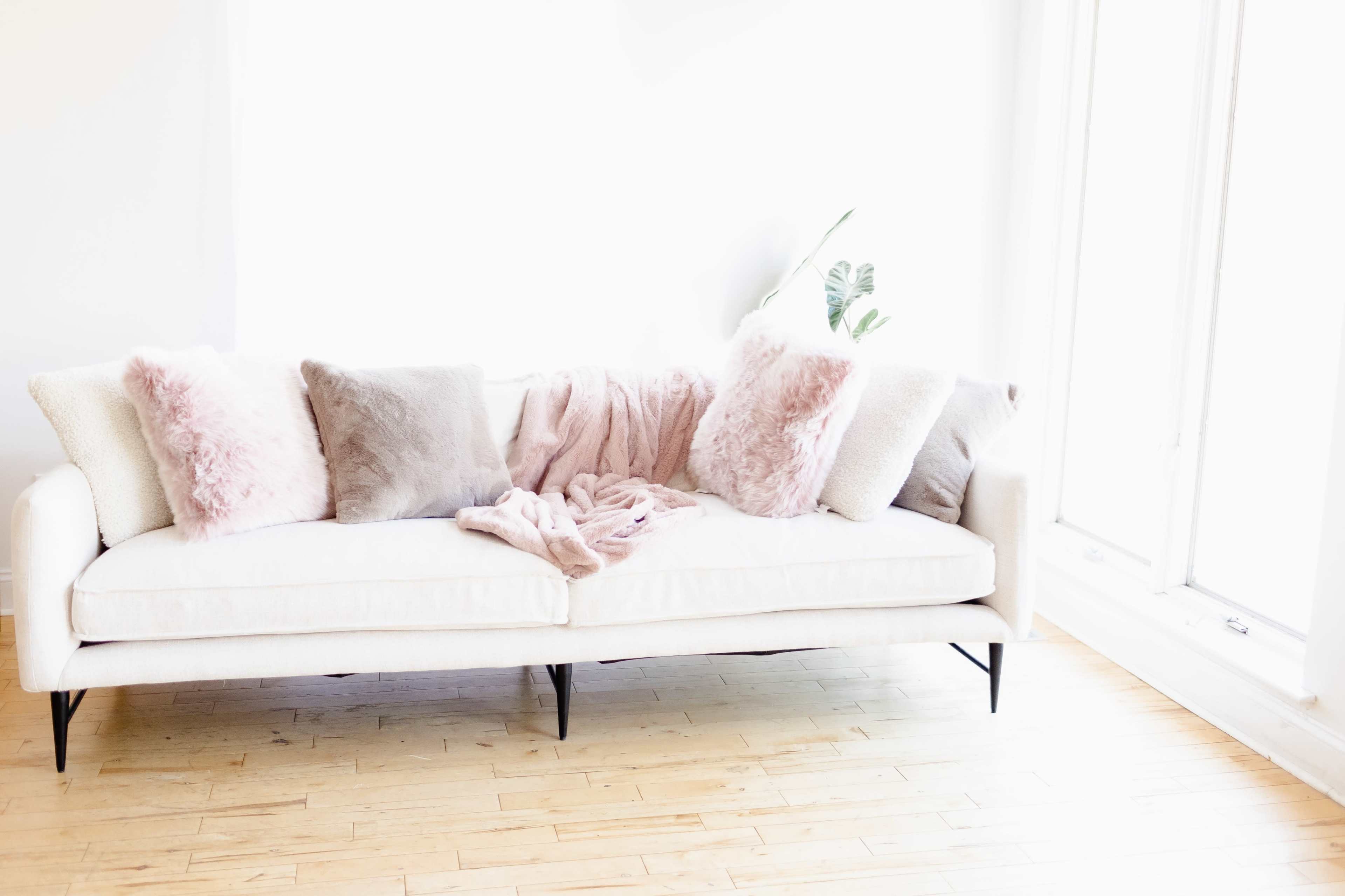 A light-colored sofa with plush pillows and a pink throw blanket is positioned near large windows on a wooden floor.