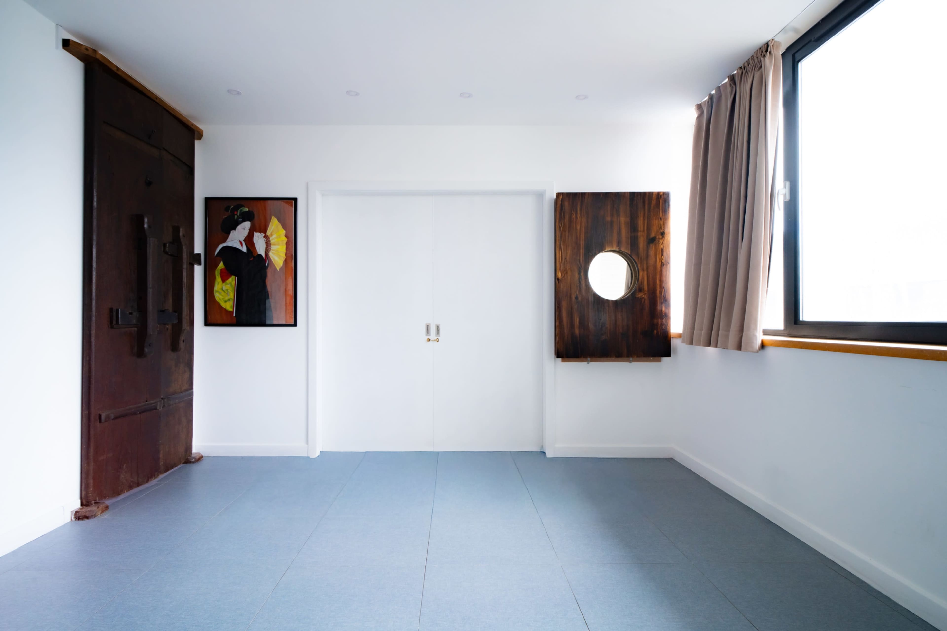 The room features a large white wall with double doors, a round window, a wooden wall panel, and framed artwork on the left.