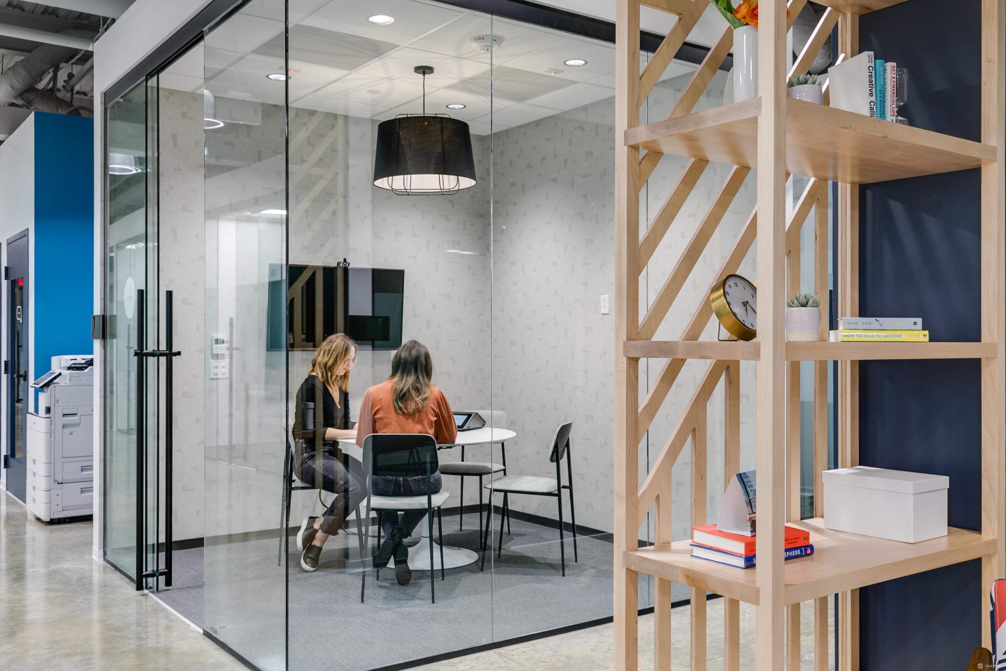 Two people are seated at a round table in a glass-walled meeting room, with a bookshelf and printer visible in the foreground.