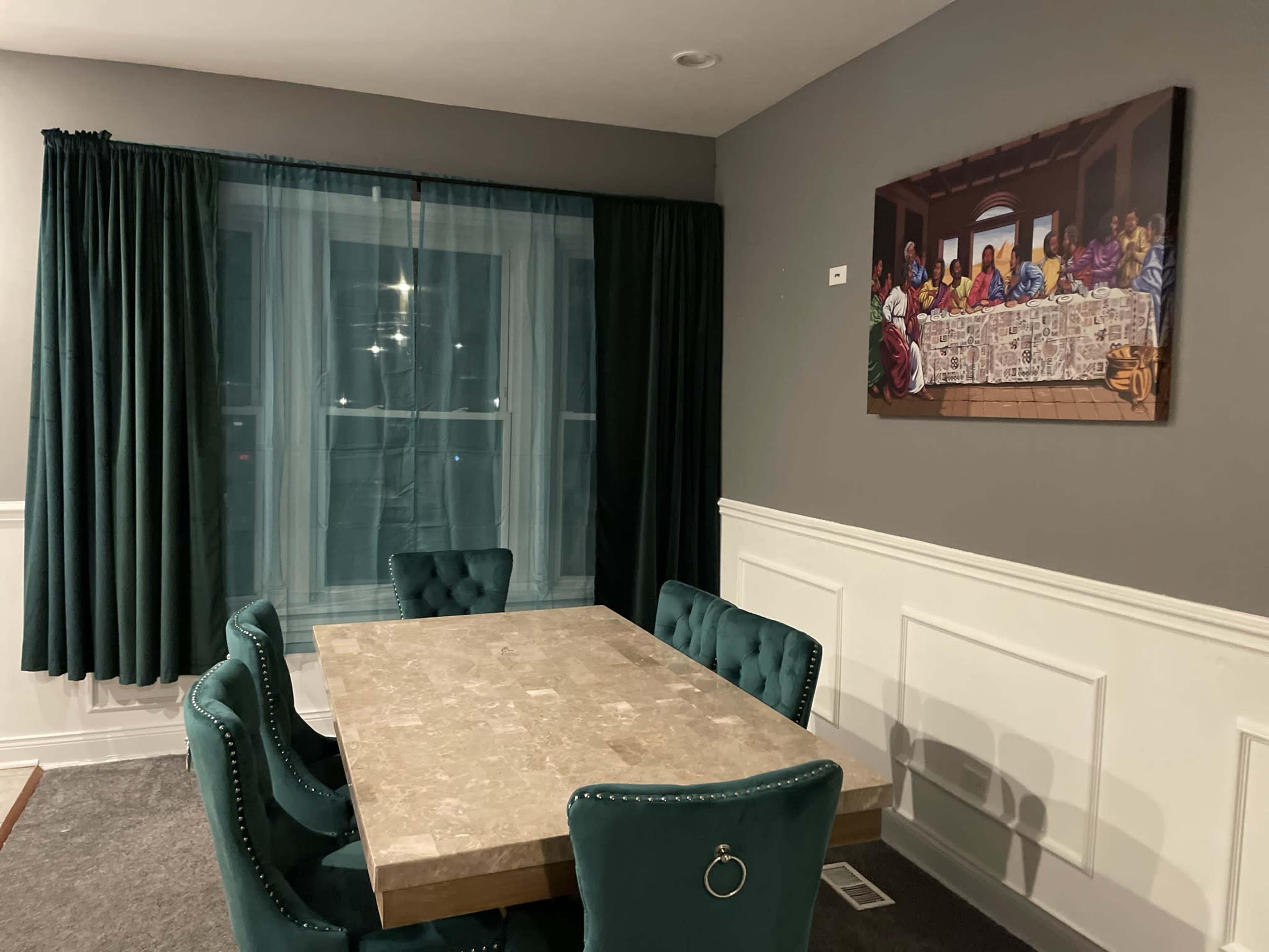 A dining room features a stone table surrounded by upholstered chairs and a large window with dark curtains, while a painting hangs on the wall depicting a group gathered around a long table.