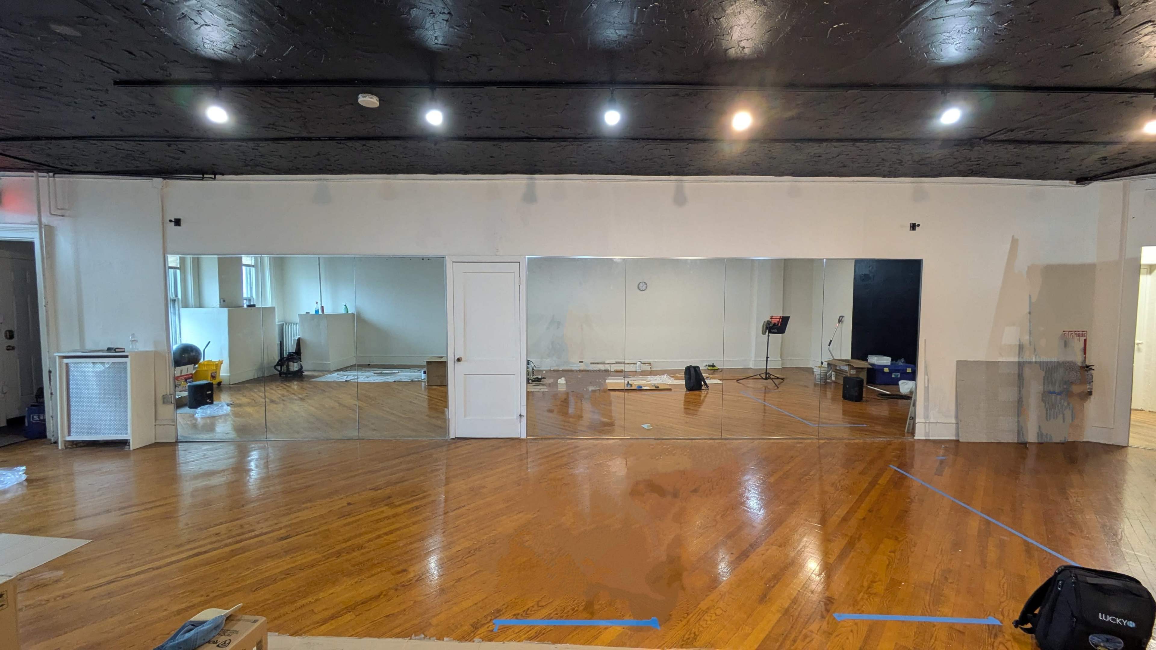 The image shows a spacious room with wooden floors, large mirrors on one wall, and various equipment scattered throughout.