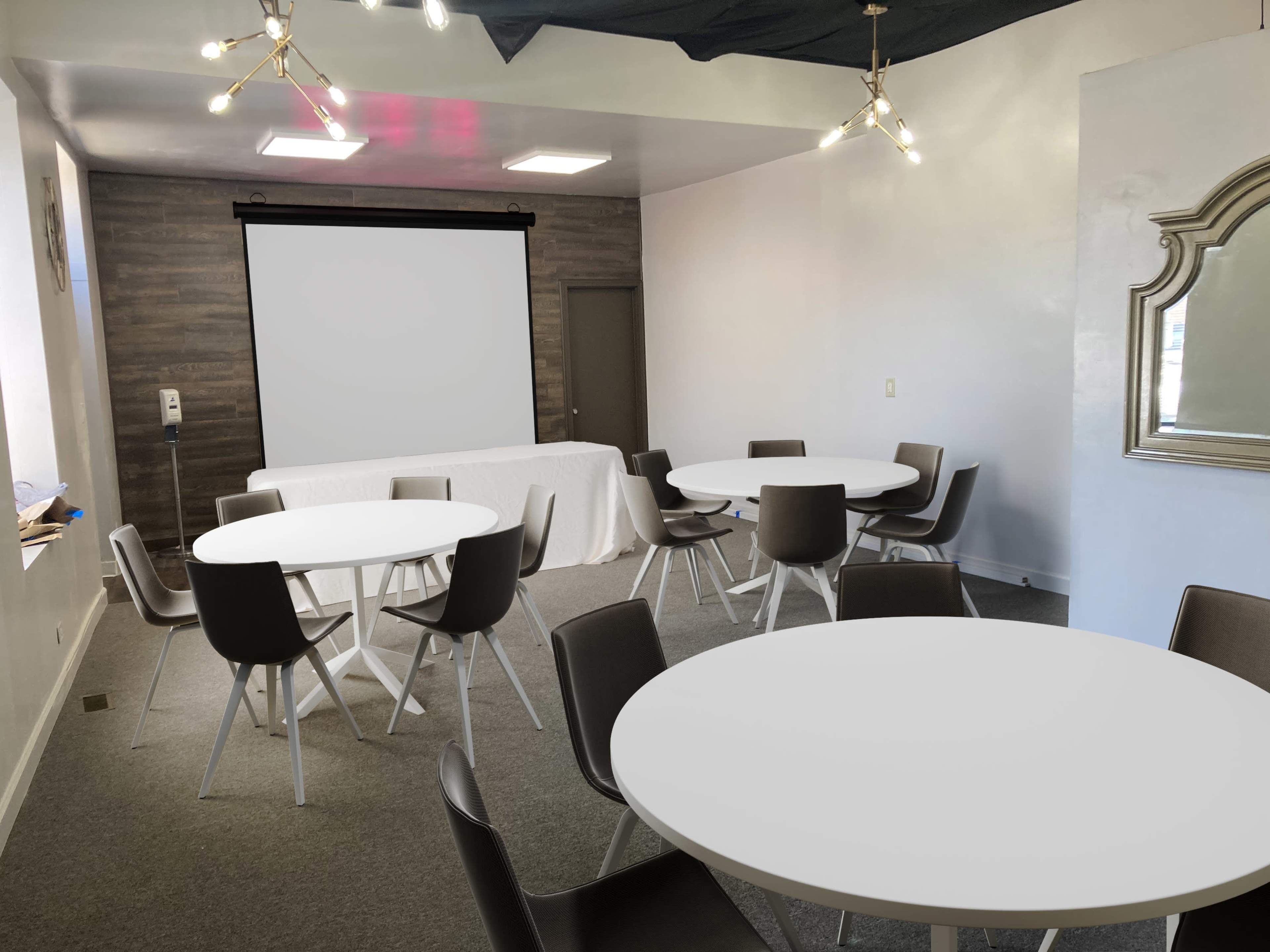 A modern meeting room with round tables and chairs, a projector screen on the wall, and a decorative mirror.