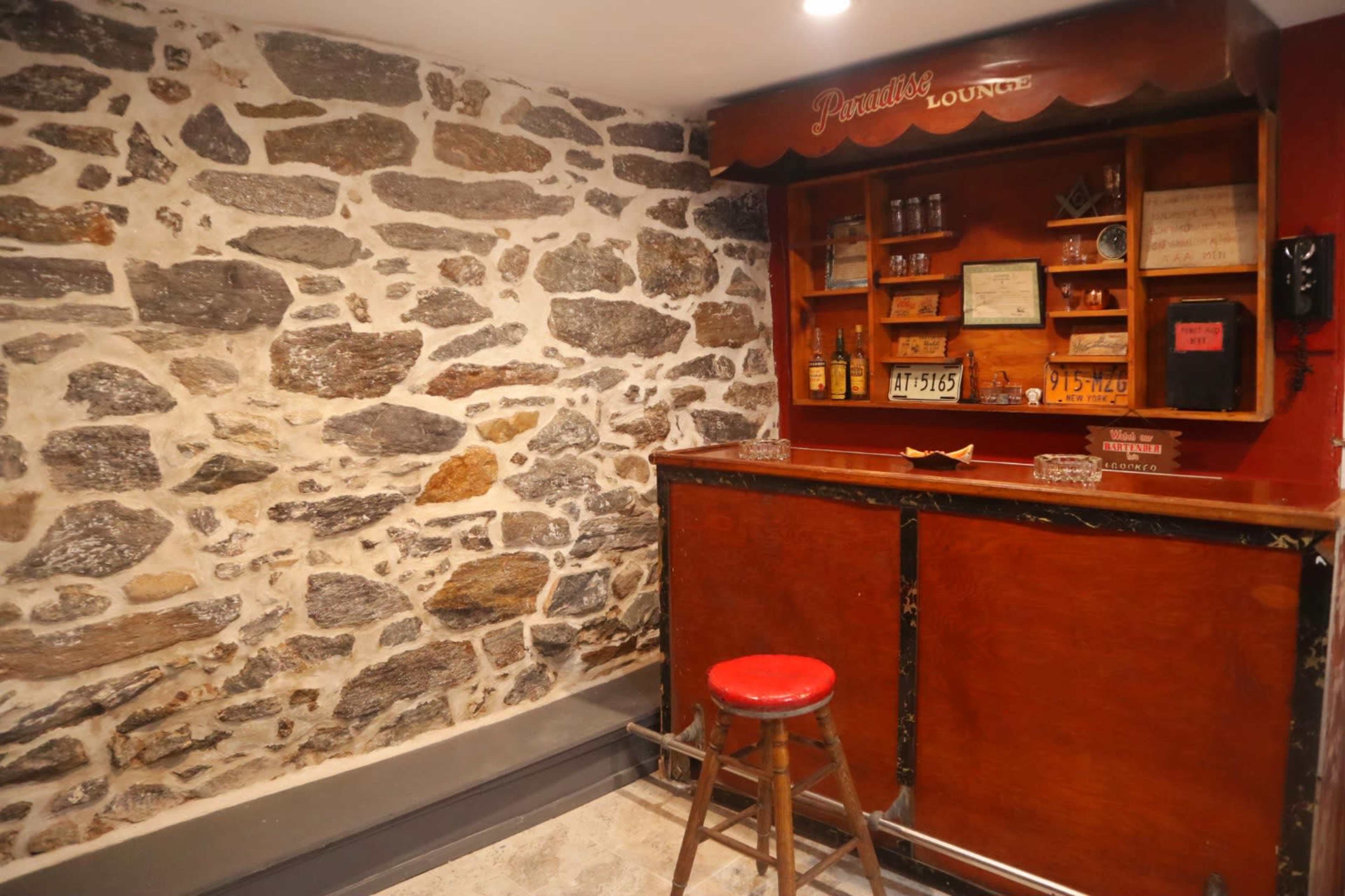 A small bar area with a wooden counter and a red stool, situated against a backdrop of a rustic stone wall.