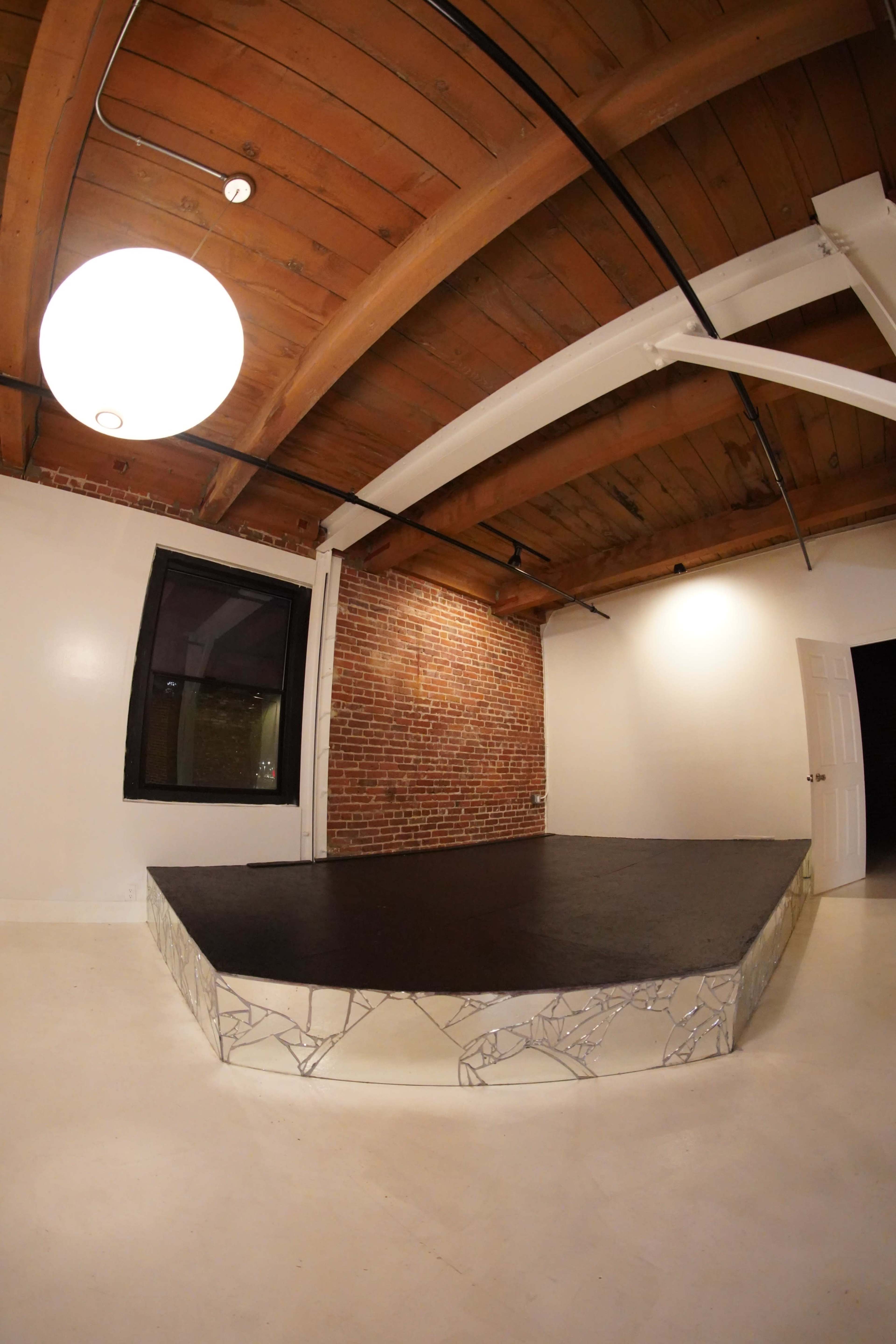 The image shows an interior space with exposed wooden beams, a circular light fixture, a brick wall, and a raised platform with a mirrored base.