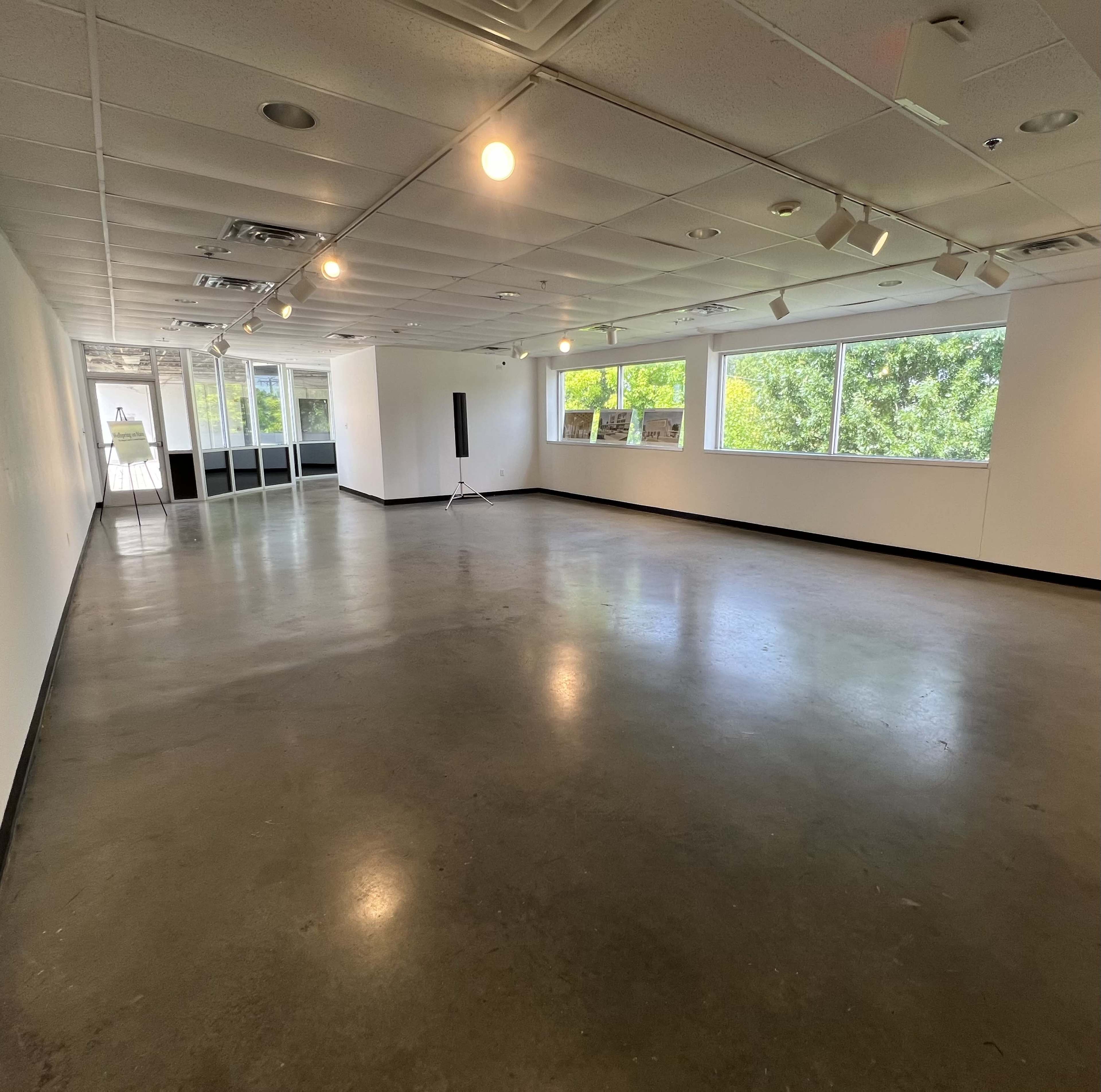 The image shows an empty, well-lit gallery space with large windows and polished concrete flooring.