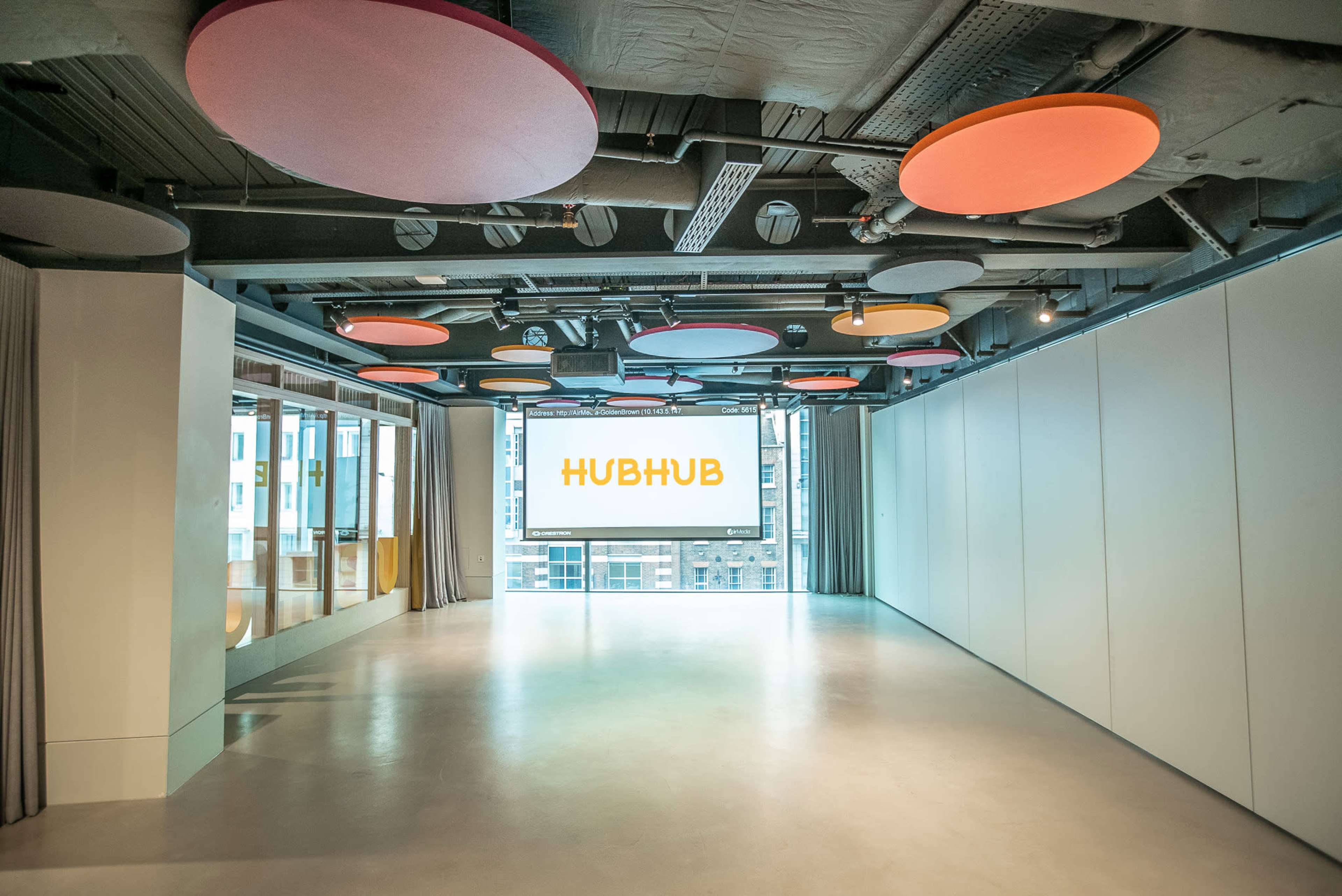 The image shows a spacious, well-lit room with colorful circular acoustic panels on the ceiling and a large screen displaying the word "HUBHUB."