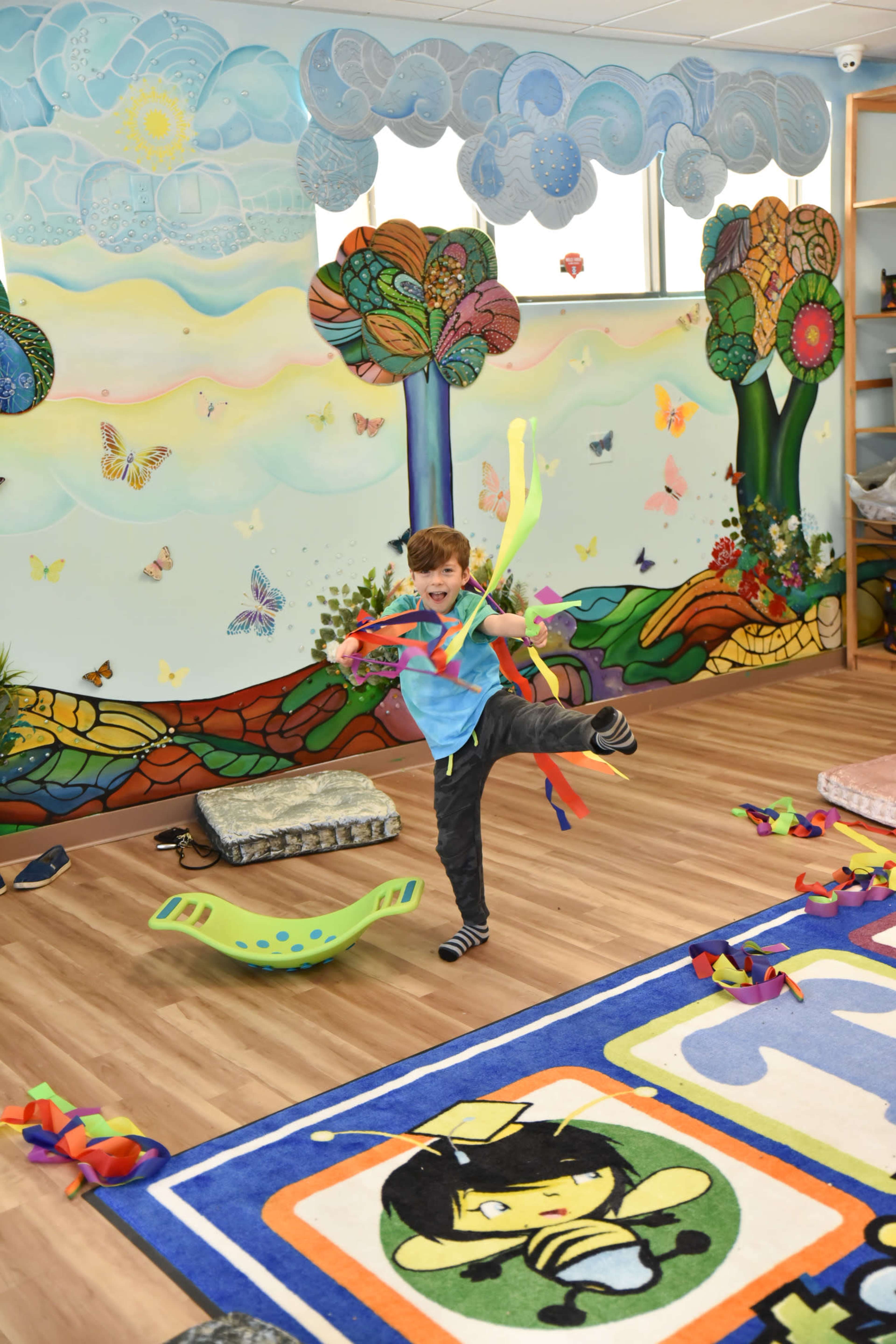 A child performs a playful pose with colorful ribbons in a brightly decorated classroom featuring whimsical murals and a variety of soft play items.
