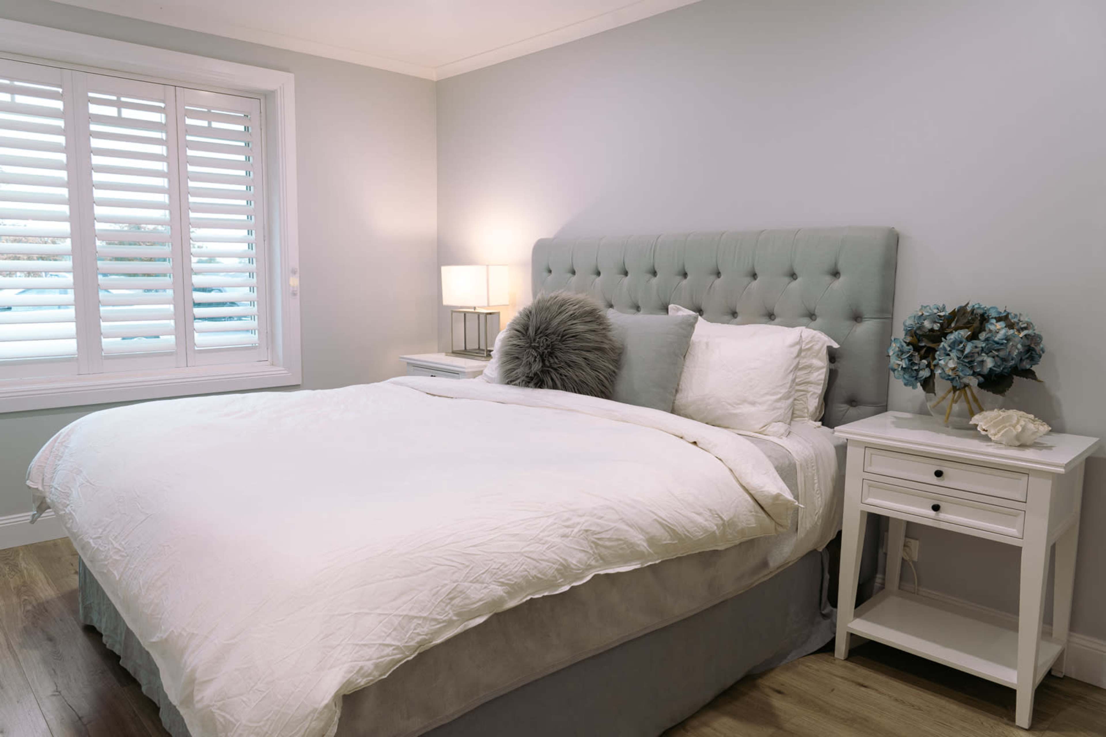 The image shows a neatly made bed with a light-colored duvet and decorative pillows in a softly lit bedroom featuring a nightstand and a window with shutters.