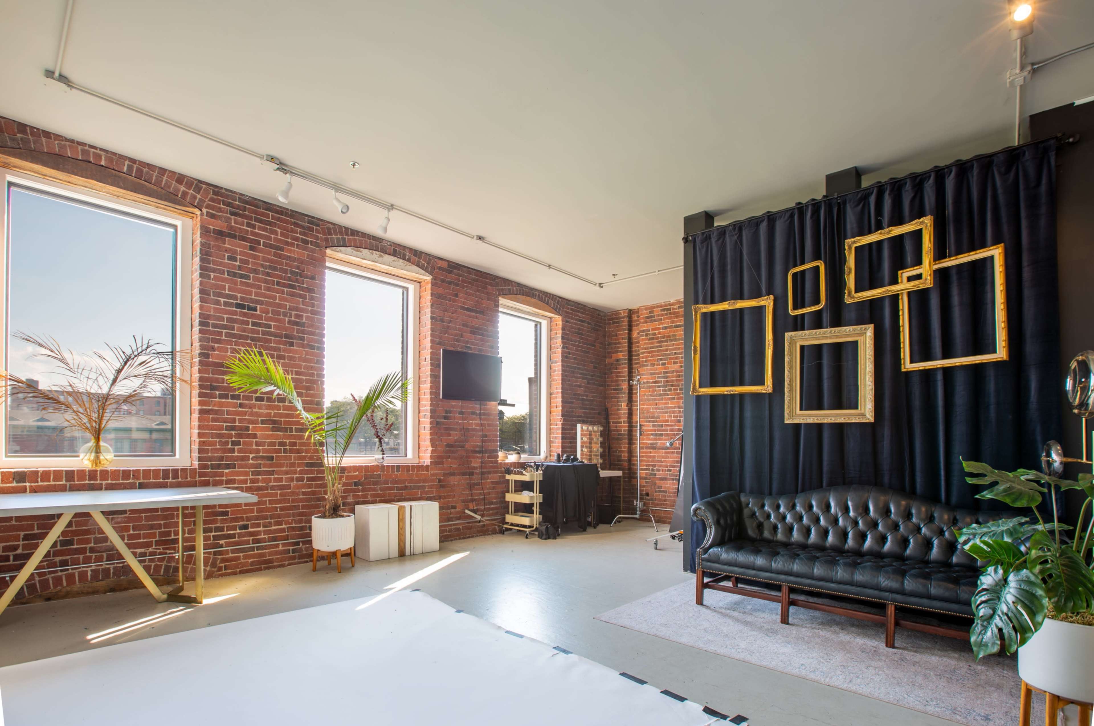 The image shows a modern interior with exposed brick walls, large windows, a leather sofa, and a dark curtain featuring empty picture frames.