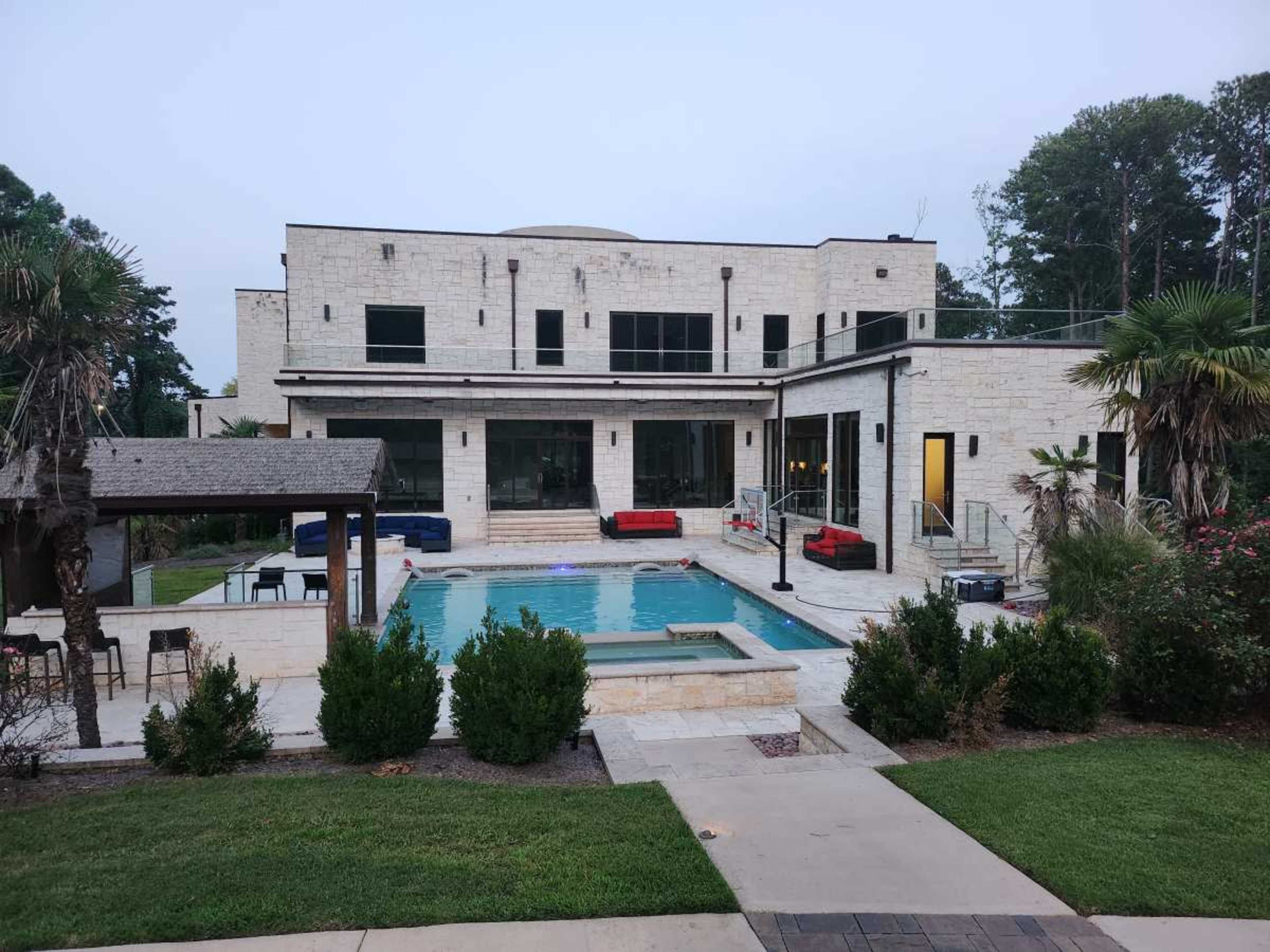 The image shows a modern two-story house with a swimming pool and a patio area surrounded by palm trees and neatly landscaped greenery.