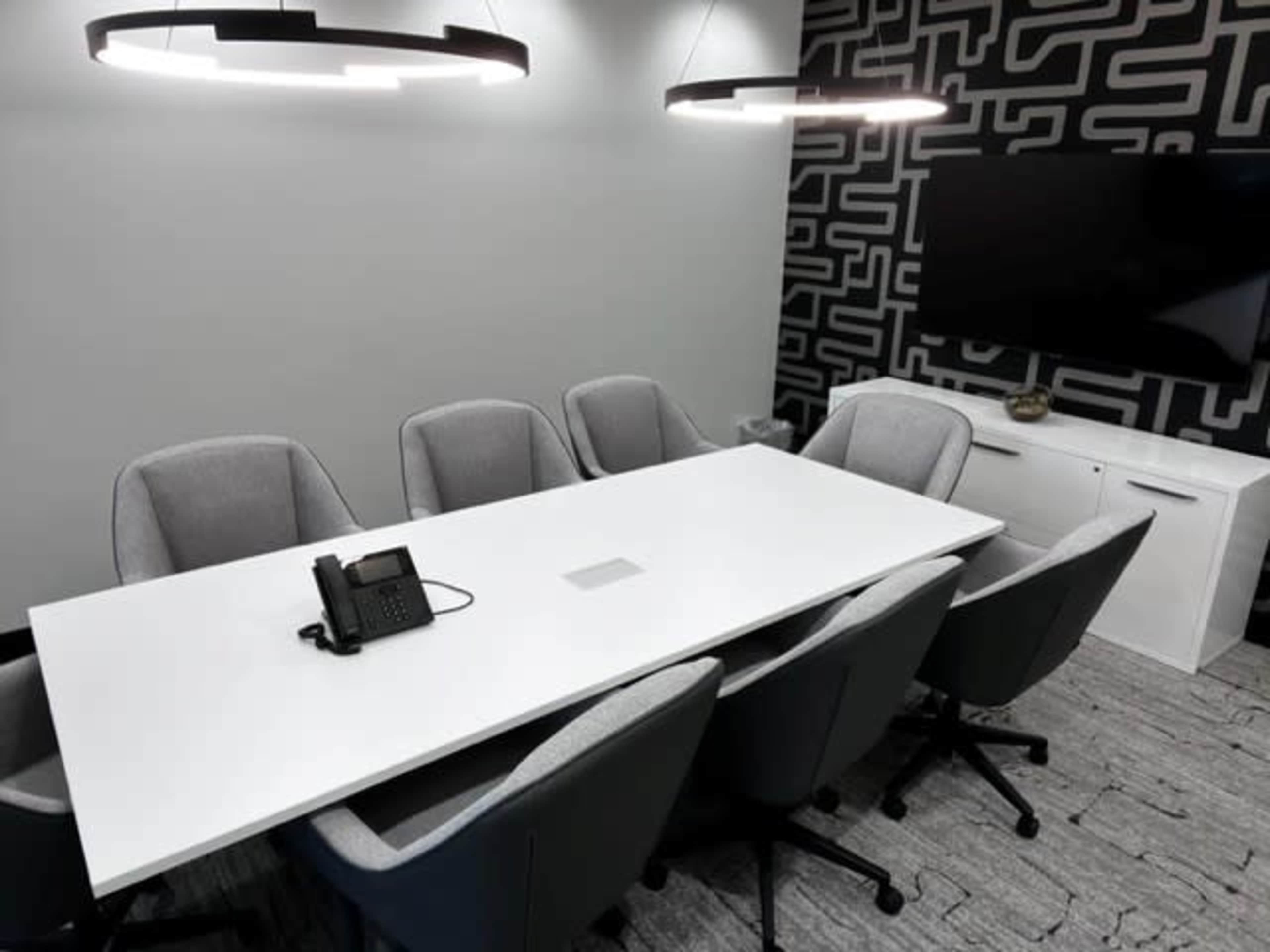 A modern conference room with a long white table, gray chairs, a phone on the table, and a television mounted on the wall.