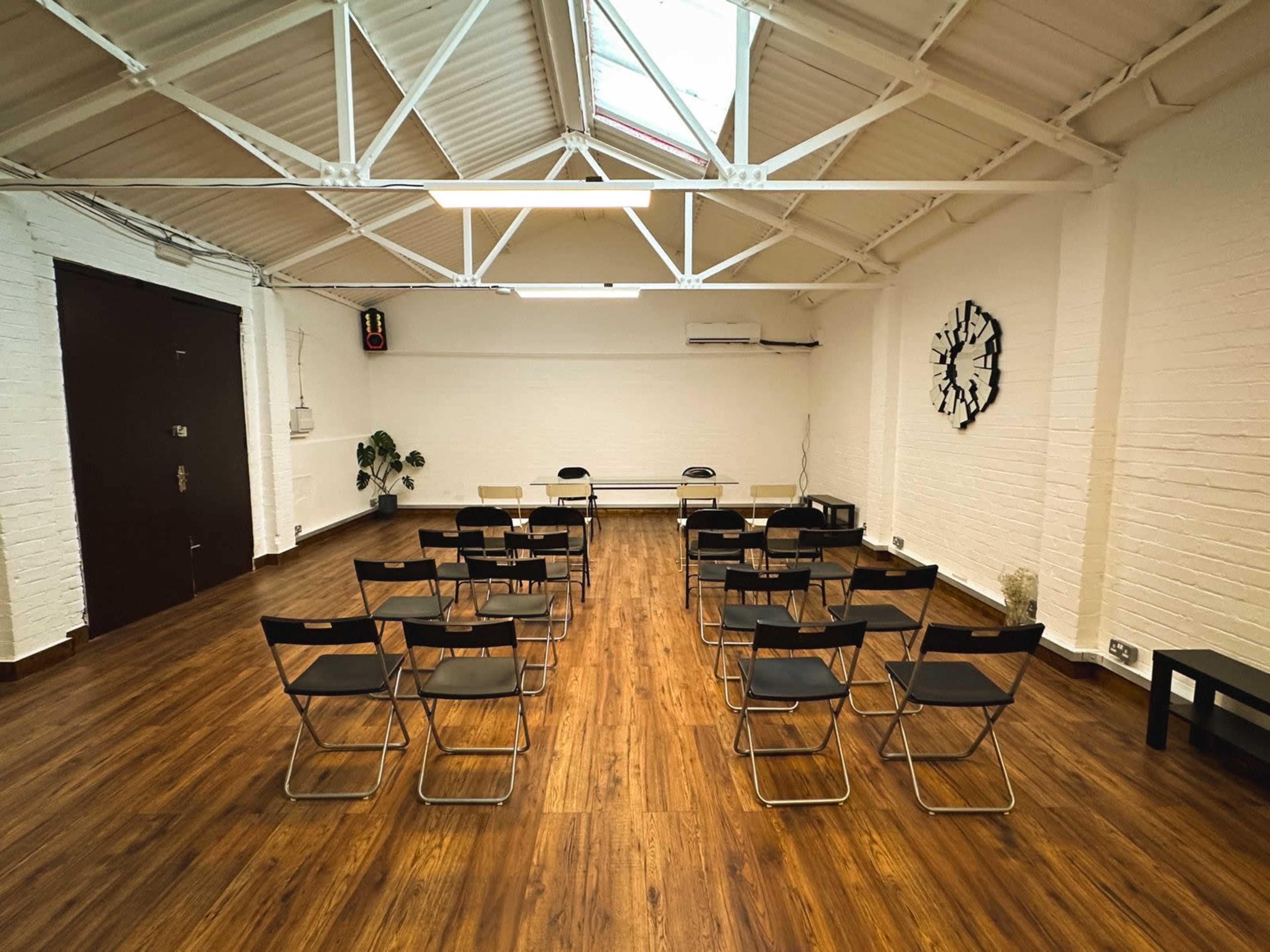 The image shows a spacious room with wooden flooring, featuring several rows of black folding chairs facing a small table and two additional chairs at the front.