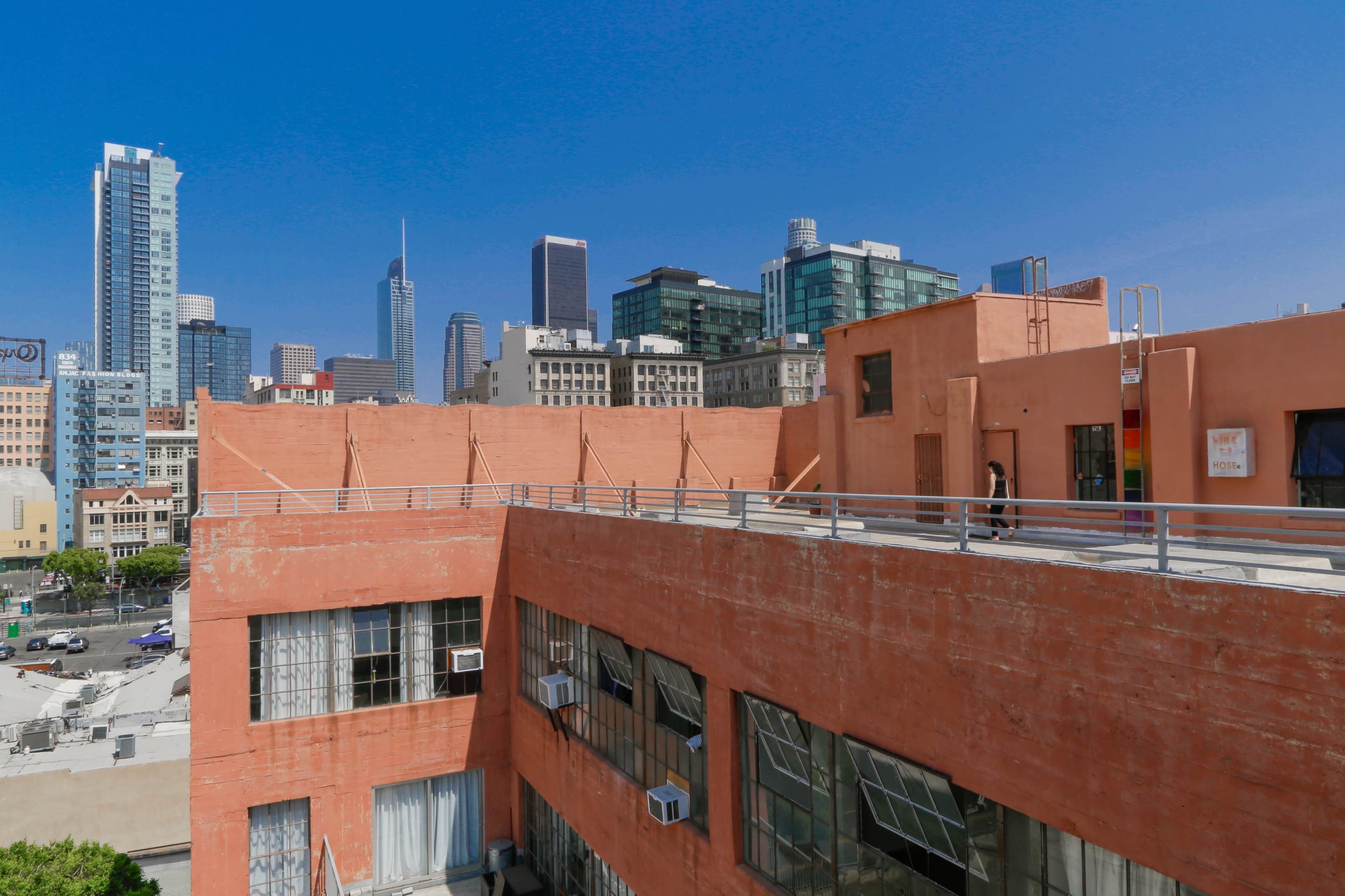 The image shows a rooftop view of a reddish-orange building with a railing, overlooking a city skyline with modern skyscrapers and clear blue skies.